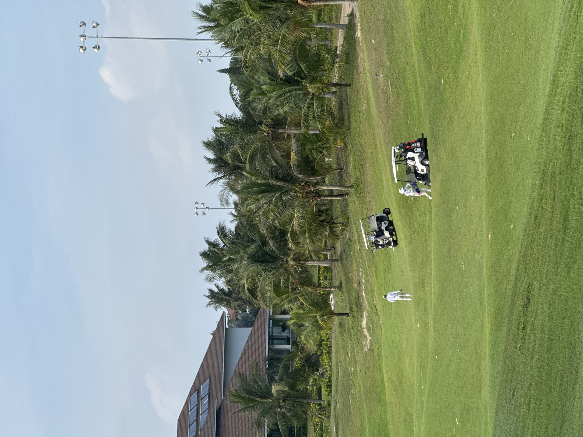 The image depicts a golf course in Phú Thủy, Vietnam, with lush green fairways and palm trees. In the foreground, a golfer in white attire is walking away from the camera, with a golf cart positioned to their right. Further in the background, another golf cart is visible with two golfers standing beside it. Beyond the golf course, a large building with solar panels on its roof and a cluster of palm trees can be seen. The sky is bright blue with scattered clouds, and tall light poles are present in the distance.