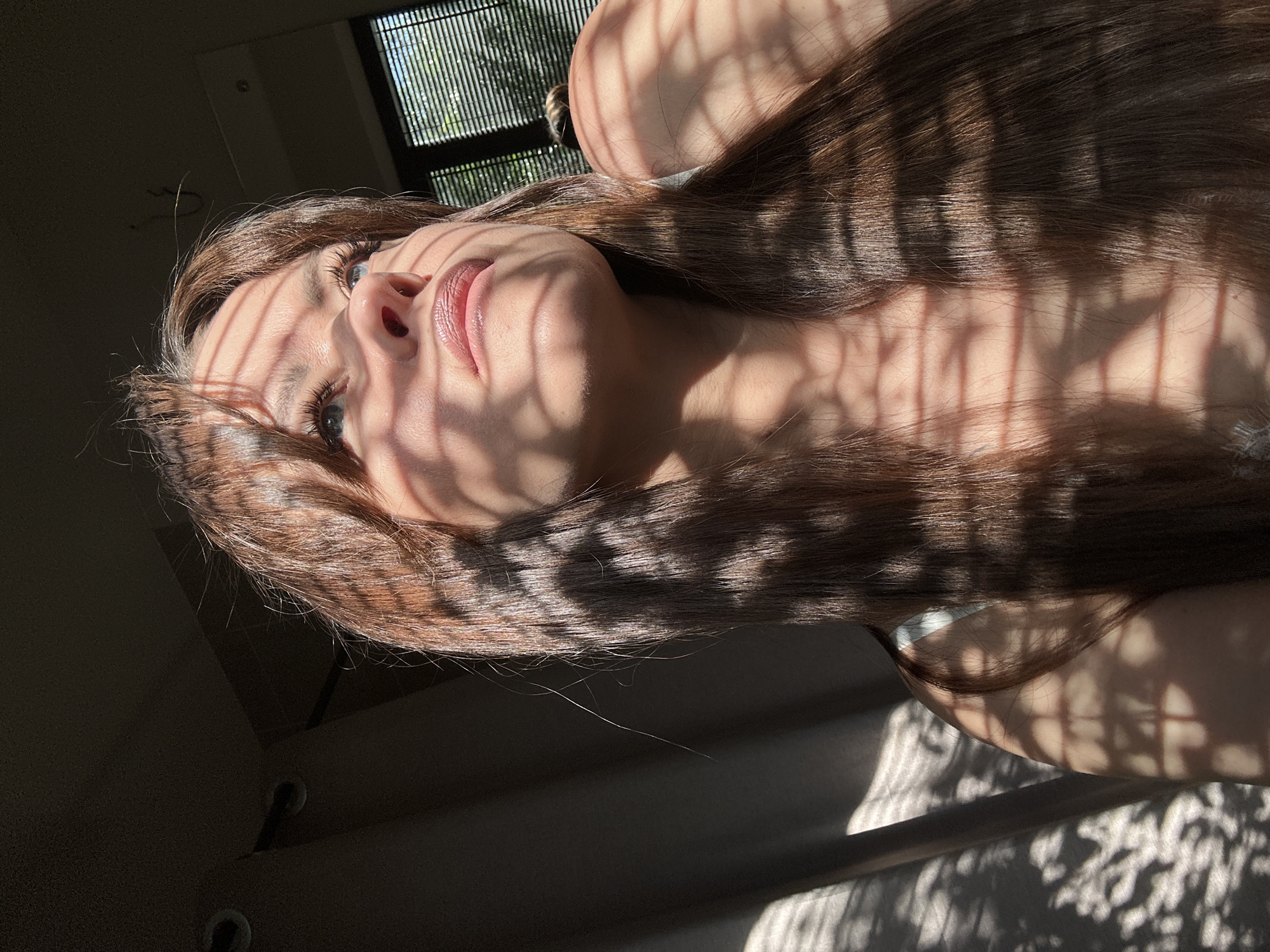 This is a vertically oriented, close-up media file featuring a young woman lying down and looking upwards. She has long, dark brown hair, which is partially illuminated by sunlight casting striped shadows across her face and body. Her skin appears smooth and lightly tanned, and her lips are slightly parted and glossy. One eye is clearly visible, showing a blue iris and long eyelashes, while the other is mostly obscured by hair.
The setting appears to be indoors, with a window featuring horizontal blinds visible in the upper left corner, suggesting it is daytime and sunny outside. The light filtering through the blinds creates the distinct shadow patterns on the woman. The background is dark and somewhat blurry, with the suggestion of a room. There are no visible objects of particular importance, apart from the implied window and blinds. The woman's expression seems contemplative or serene, perhaps enjoying the warm sunlight. The shadows create a dramatic and artistic effect, highlighting her features and hair. The overall mood is peaceful and intimate. No text is visible in the media file.