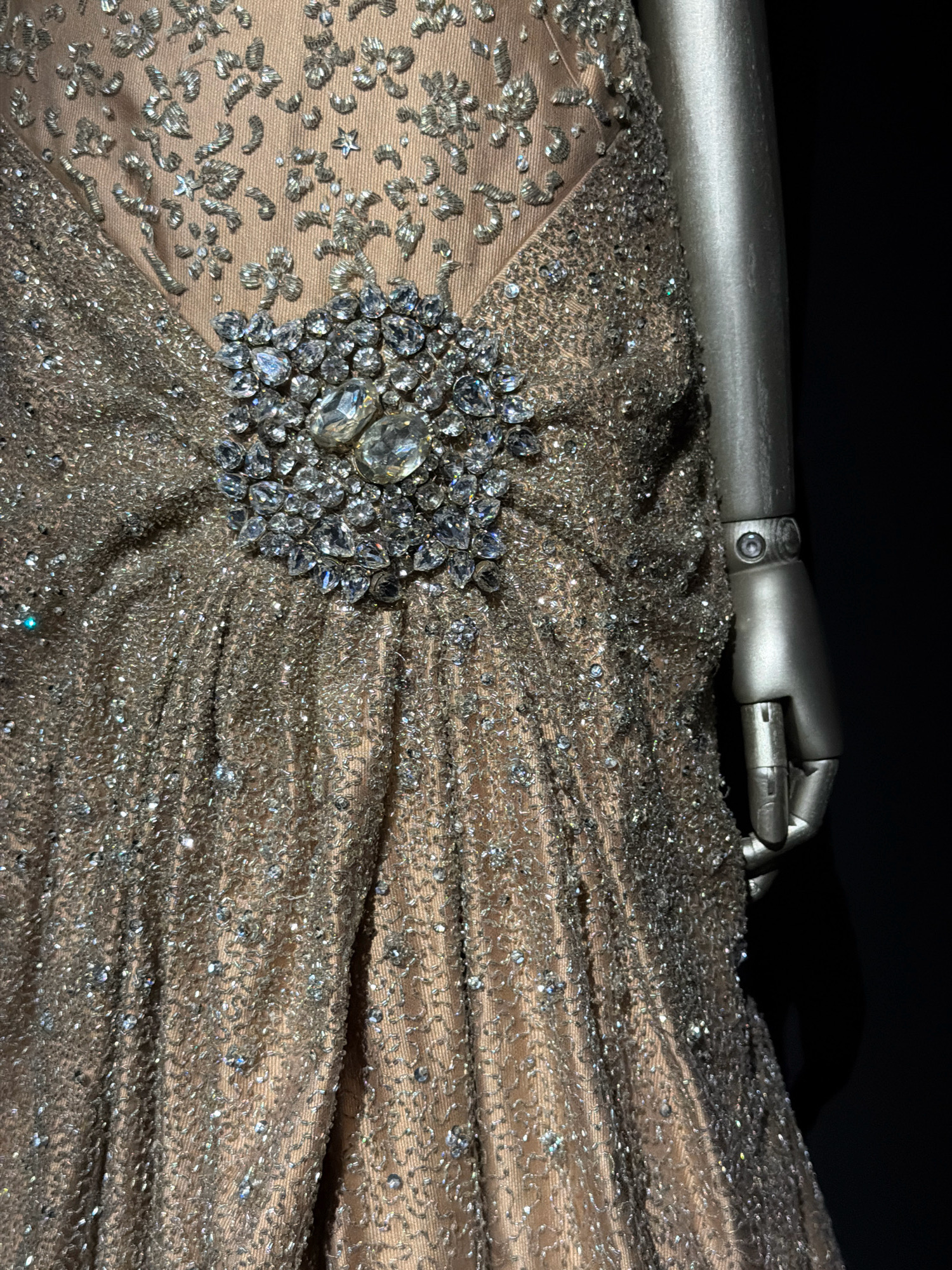 The image displays a close-up view of a heavily embellished gown on a mannequin, suggesting a museum or exhibition setting. The gown features intricate beading and sequin work in silver and clear gemstones, primarily concentrated around the bodice and cascading down the fabric. The fabric itself appears to be a light brown or taupe silk with vertical ruching. A prominent brooch, composed of large clear gemstones and smaller surrounding stones, is affixed to the center of the bodice. The right arm of a silver mannequin is visible, extending from the side of the frame, with its hand positioned near the hip of the gown. The background is predominantly black, with a slight sheen on the mannequin's surface and a subtle light source highlighting the gown's details. There is no indication of the city of Milan, Italy, within the image or its context.