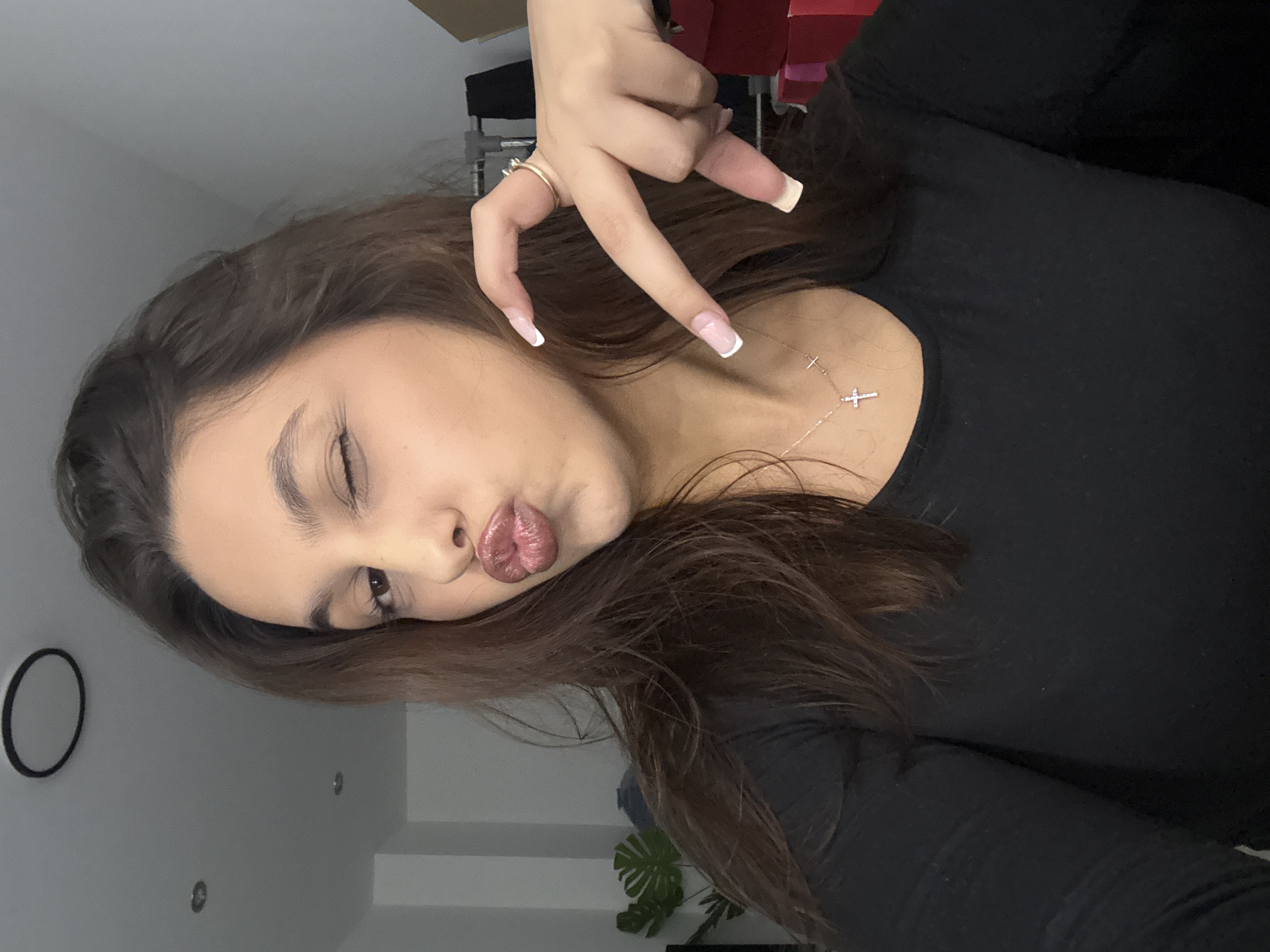 A young woman is captured in a casual selfie, holding her phone to take the picture, evident by the upward angle and pose. She has long, dark brown hair and is wearing a black long-sleeved top. Her head is tilted, and she sports a playful expression with her left eye winking and her lips puckered into a "kiss face," highlighted by glossy, reddish-pink lipstick. Her eyebrows are well-groomed.
She holds her right hand up, making a "peace sign" gesture with her index and middle fingers extended, while her ring finger and pinky are curled. Her long, squared nails feature a classic white-tipped French manicure. A simple gold band ring is visible on her right ring finger. Around her neck, she wears a delicate, silver-toned necklace adorned with two small cross pendants.
The background is softly blurred but appears to be an indoor setting with light-colored walls. A plain black circular object is mounted on the wall to the left, and a green plant with large leaves, possibly a Monstera, is visible in the lower right. Some indistinct dark furniture or equipment parts are also present in the background. There is no visible text in the image. The overall mood conveyed is casual, playful, and confident. This image was taken in Mönchengladbach, Germany.