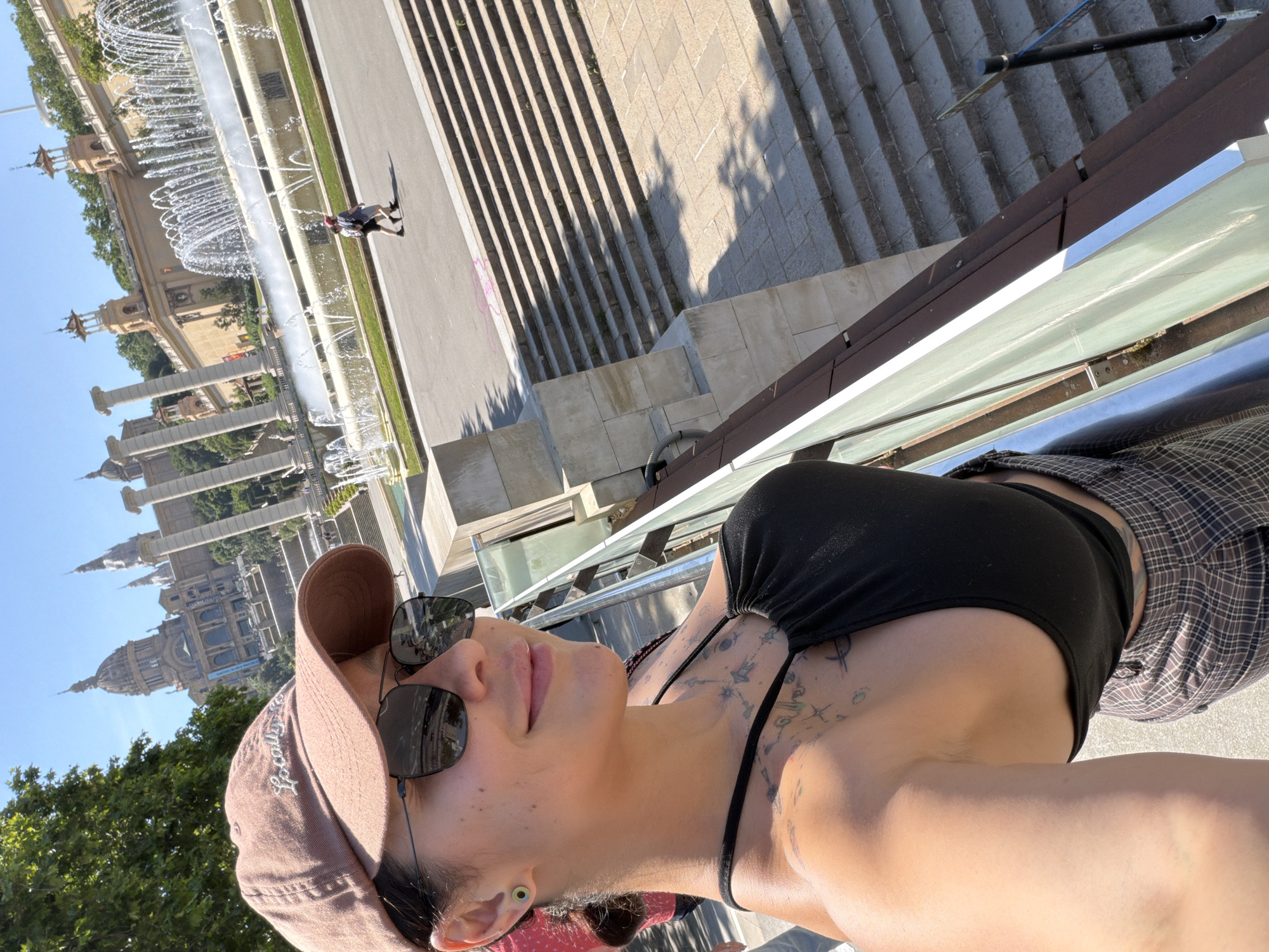 A photo of a woman taken on a sunny day in Barcelona, Spain, with the Magic Fountain of Montjuïc and the National Palace in the background. The woman, wearing a brown baseball cap and sunglasses, stands against a glass barrier. She wears a black top and a plaid skirt. Steps are behind the barrier.