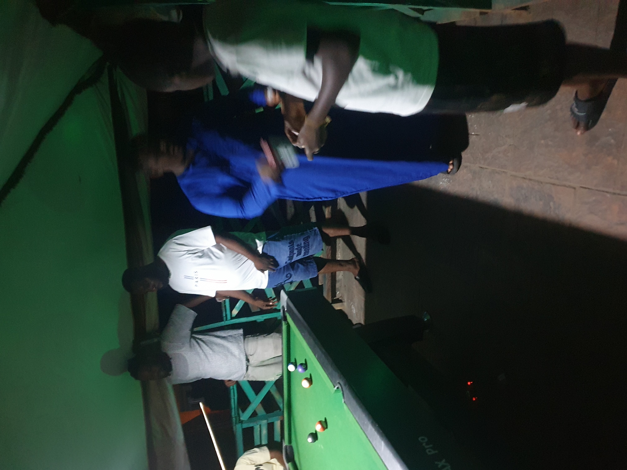 The image depicts a group of individuals gathered around a pool table at night, possibly in Lokoja, Nigeria. The scene is dimly lit, with green overhead lighting casting a shadow on the surroundings.
In the foreground, a pool table with several balls, including a cue ball, a striped ball, and a solid red and orange ball, is visible. A person in a white t-shirt and dark shorts is standing to the right of the table, holding a pool cue. Further back, another person in a bright blue robe is interacting with a person holding a small item. To the left, two men are standing near the pool table. One is wearing a striped shirt and light-colored shorts, while the other is wearing a white t-shirt. A pool cue is resting on the edge of the table. The environment appears to be an outdoor or semi-outdoor area with a wooden railing in the background. The ground is paved with what looks like concrete or stone tiles. The overall atmosphere suggests a casual social gathering.