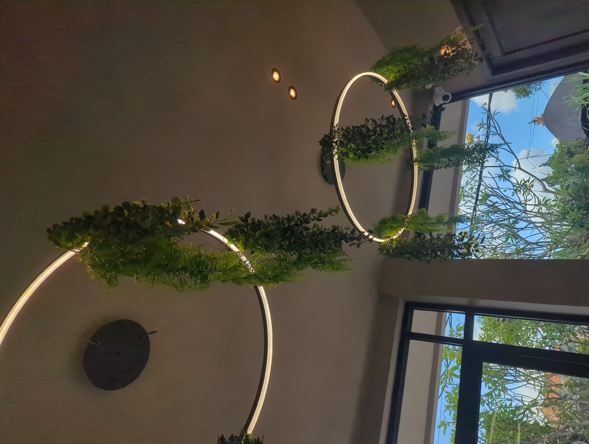 A low-angle shot indoors reveals circular LED light fixtures adorned with green foliage. The room is dimly lit, with the walls being a neutral tone. Two small, circular lights are embedded in the ceiling. Through large windows framed in black, a view of the outside is visible, showcasing trees and a blue sky with clouds. A security camera is mounted near one of the windows. A circular object is affixed to the wall near the bottom left corner of the image. The image location is Mengwi, Indonesia.