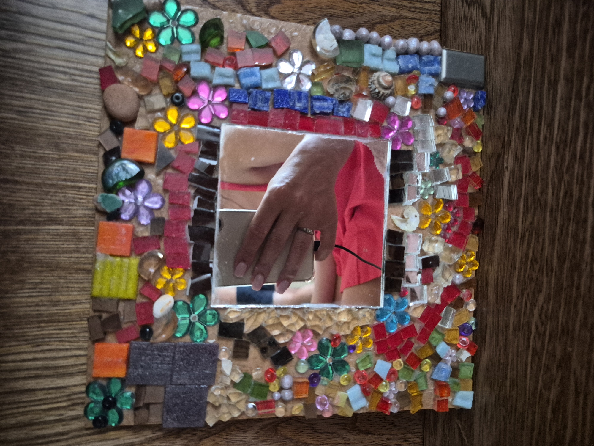 A handmade mosaic mirror frame is displayed on a wooden surface. The frame is rectangular and covered with a colorful assortment of small glass tiles, beads, and other decorative elements, including small shell fragments and flower-shaped embellishments. The central part of the frame is a mirror, which reflects a hand holding a smartphone, suggesting the photo was taken by the person in the reflection. The hand is adorned with a ring on the index finger. The person's torso is visible in the reflection, wearing a bright red garment. The background of the mosaic is a light brown base, and the entire frame rests on a surface with a distinct wood grain pattern, likely a table or shelf. The lighting suggests an indoor setting.