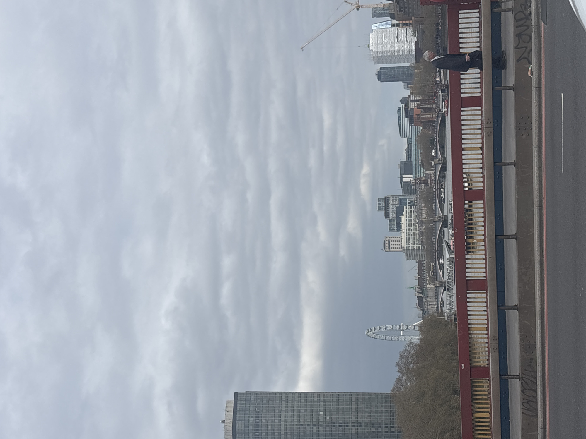 The image depicts a view of London, United Kingdom, possibly from a bridge. The sky is overcast. The London Eye is visible along with other buildings and a construction crane. A person wearing a dark suit is standing beside a painted metal railing. The road surface is gray with a white dividing line. Graffiti is present on the railing and road surface.