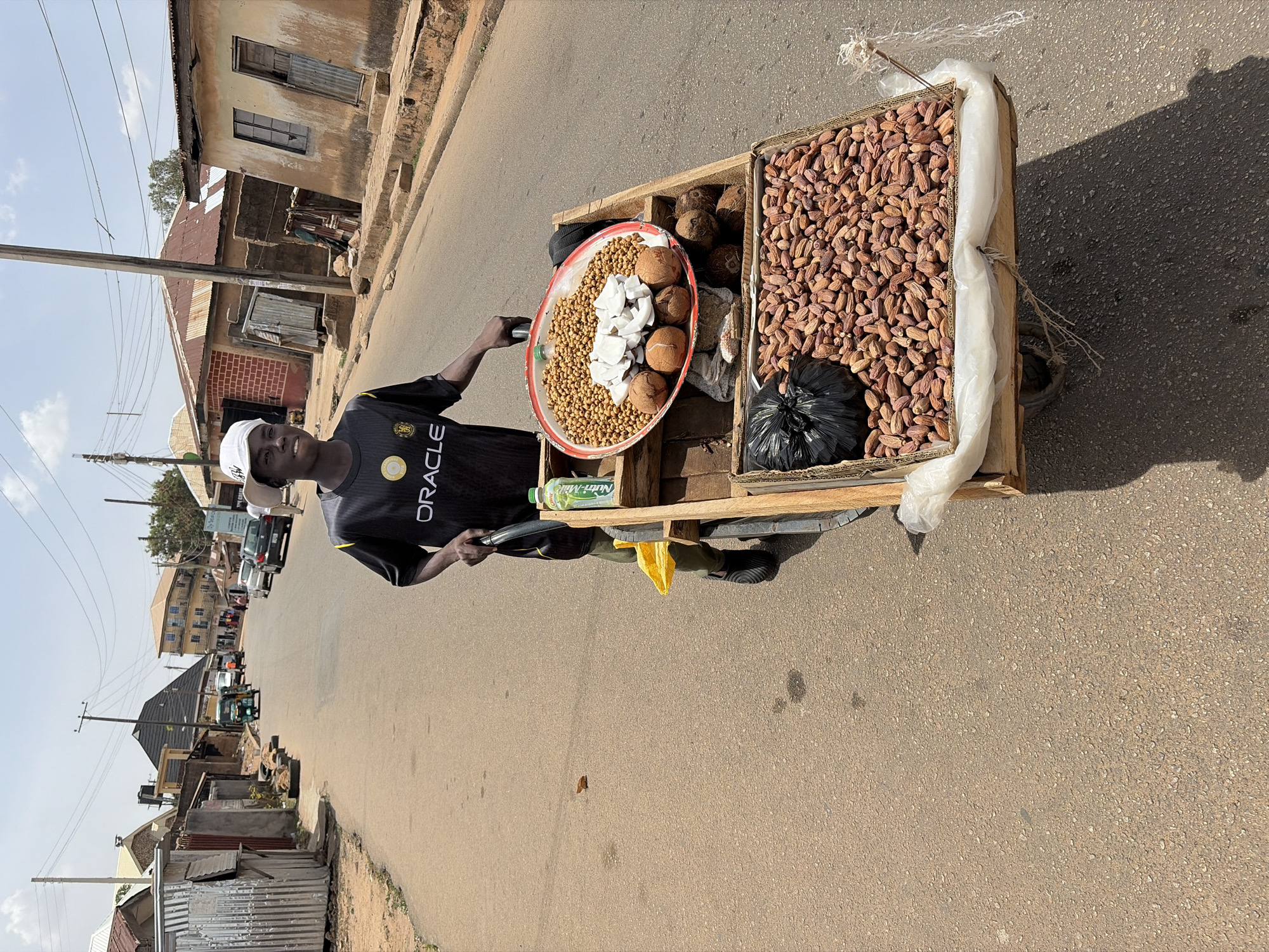A young street vendor is captured standing beside his pushcart on a paved road in Jos, Nigeria, under bright, clear daylight. The man, facing the camera with a smile, wears a black t-shirt with "ORACLE" printed on it, a white baseball cap, and green trousers.
His wooden pushcart displays a variety of goods for sale. A large, red-rimmed metal tray on the cart holds a mound of what appear to be roasted groundnuts (peanuts), several whole coconuts, and neatly arranged white pieces of processed coconut meat. Adjacent to this, a large wooden crate is filled with numerous dates, with a black plastic bag nestled among them. A green bottle, visible beneath the tray, has "Nutri-Milk" printed on its label.
The scene depicts a typical street in a Nigerian town. In the background, modest buildings with weathered, light-colored walls and corrugated iron roofs line the road. Utility poles with overhead wires are also prominent features. The sun is high, casting distinct shadows from the cart and other objects, indicating a sunny day. The man's friendly expression suggests a welcoming presence in this everyday street market setting.