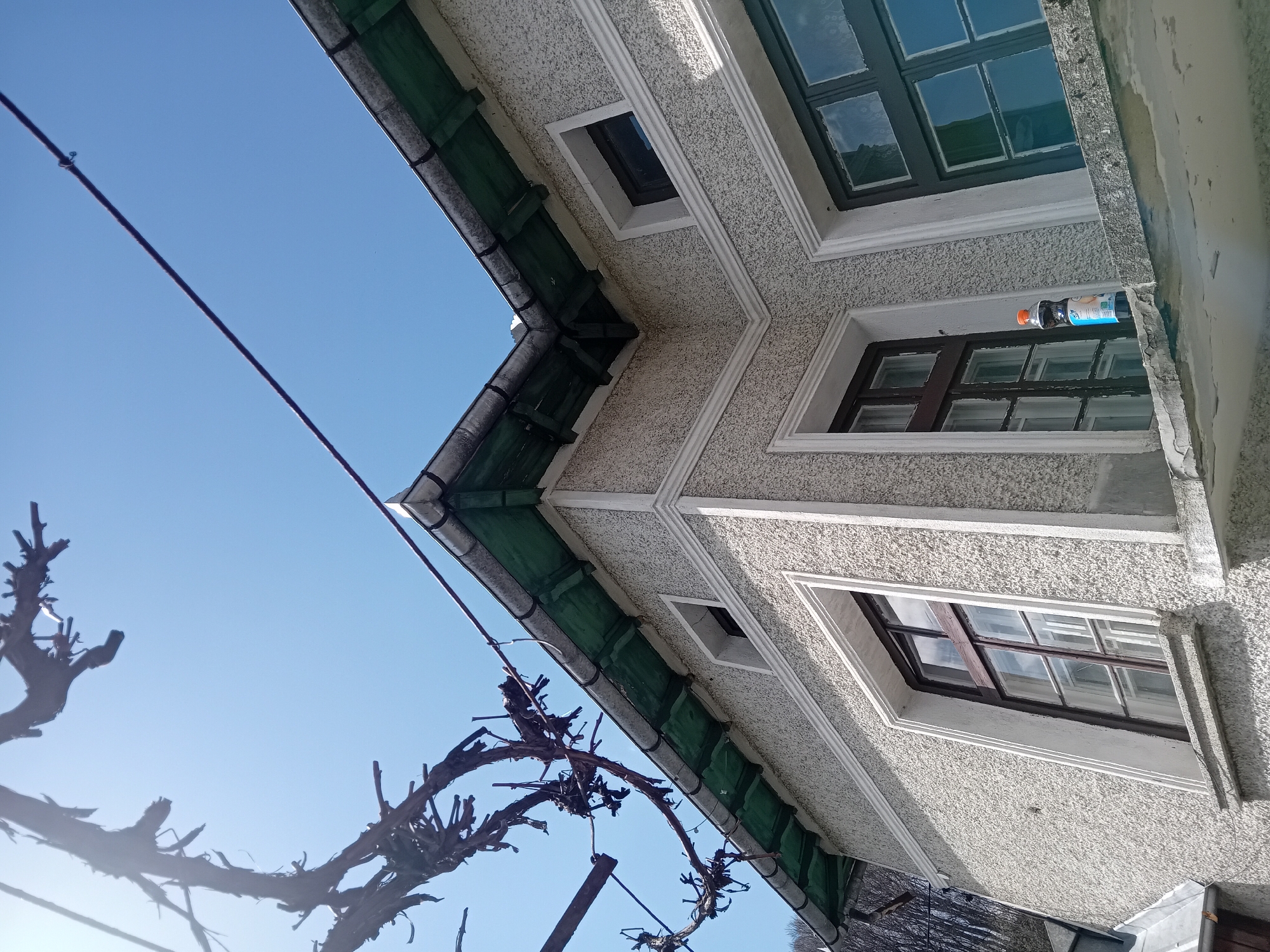 A low-angle shot depicts a building against a clear blue sky, with a tree branch in the foreground. The building has a textured grey facade, with dark green gutters and white trim around windows. Windows have a multi-pane design, and the window frames are painted brown and green. A bottle is resting on a window ledge. A dark wire stretches across the sky. The location is Unknown, Unknown.