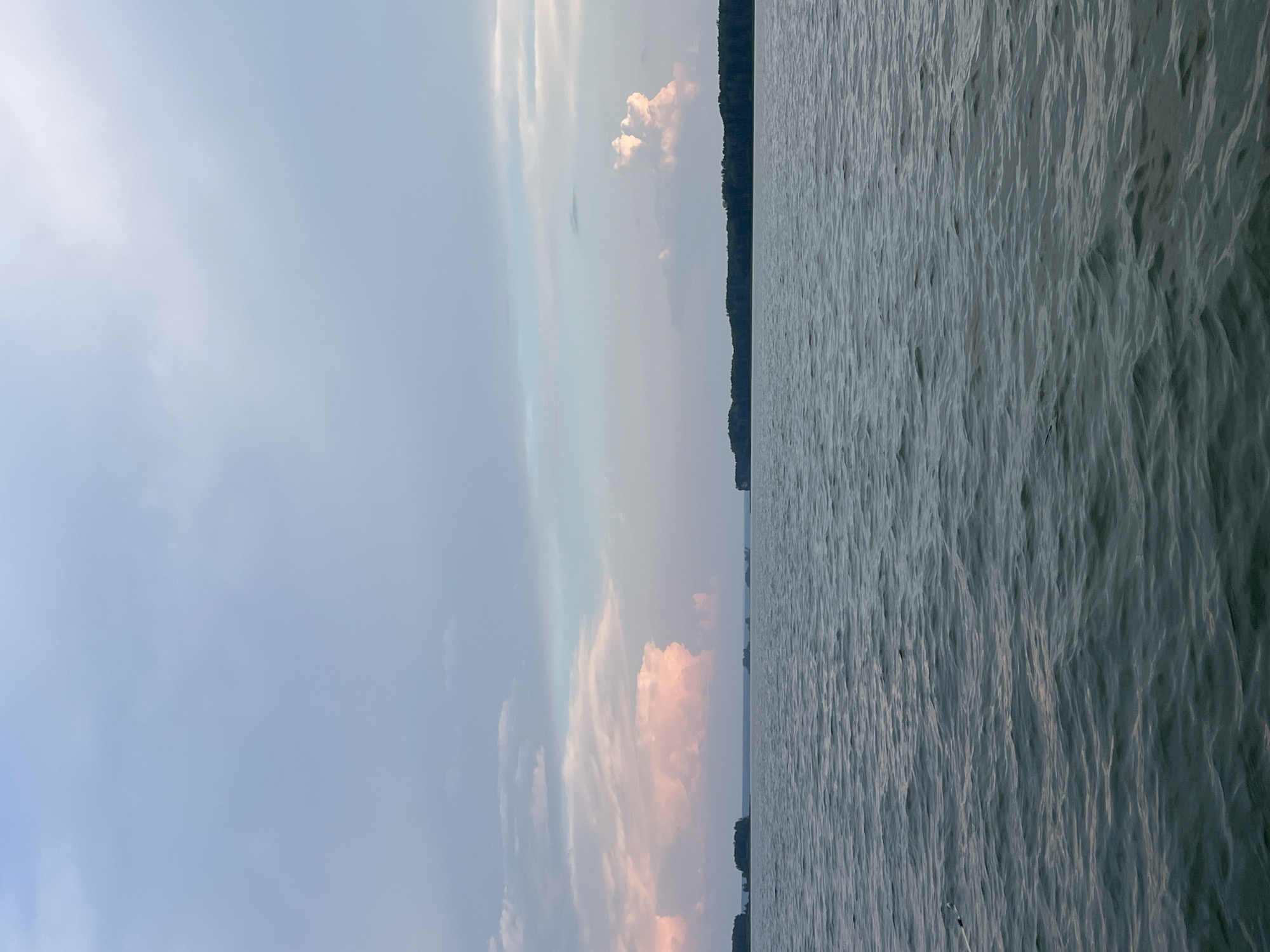 The image shows a tranquil body of water with a distant, tree-lined shore and islands. The sky above is filled with a mix of soft grey and pale blue clouds, with hints of pink in some of the formations, suggesting either sunrise or sunset. The water's surface is rippled, reflecting the muted colors of the sky. There are no people, animals, or man-made structures visible in the scene. The environment appears to be a natural landscape. The location in Novopichugovo, Russia, is not visually discernible from the image itself.
