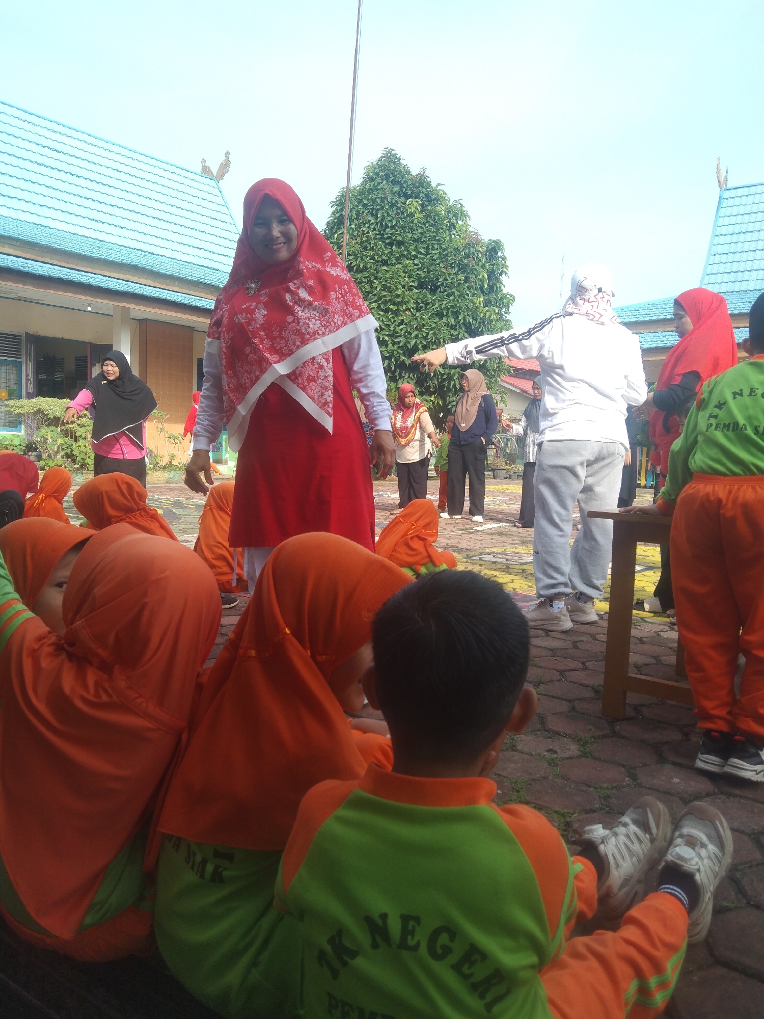 The image shows an outdoor scene at what appears to be a school in Siak Sri Indrapura, Indonesia. In the foreground, several young children wearing orange hijabs and green and orange uniforms are sitting on the ground. Behind them, a woman in a red hijab and red dress is standing and smiling at the camera. To the right, a person in a white tracksuit and grey pants is pointing to the left. Other adults and children are visible in the background, some wearing hijabs. The environment includes a building with a blue roof, trees, and a paved ground surface. The lighting suggests it is daytime.