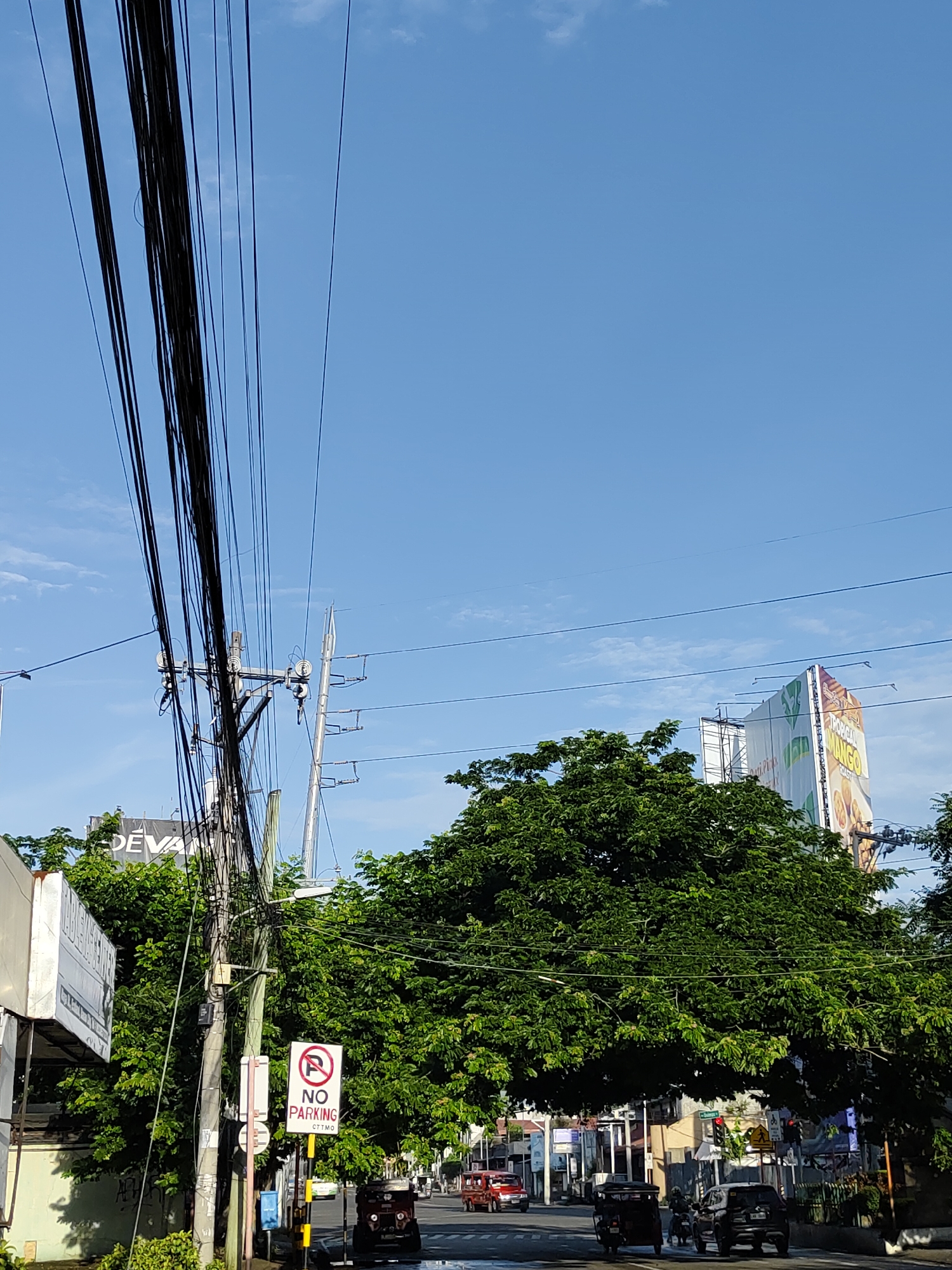 The image captures a bustling street scene in Davao City, Philippines, under a clear, bright blue sky with scattered wispy clouds. Numerous black utility wires and cables crisscross the upper left and center, originating from a wooden utility pole that dominates the left-center foreground.
Green, leafy trees with dense foliage are prominent, partially obscuring buildings and billboards in the midground and background. On the right, a large billboard is visible, featuring text that appears to read "TROPICAL MANGO" alongside an image of food. Another billboard on the left has "DEVA" or "DEVANT" partially visible.
On the road, several vehicles are present, indicating active traffic. These include a dark-colored tricycle positioned left of center, a dark SUV further to the right, and a red vehicle (possibly a jeepney) at the far left. A red jeepney is also partially visible on the far right.
A white "NO PARKING" sign with a red circle and slash is prominently displayed in the lower-center, with "CTTM O" written below it, likely referring to the local traffic management office. Other street furniture includes a small blue bin and distant traffic lights. The scene suggests a sunny day, likely morning or early afternoon, in a typical urban environment.