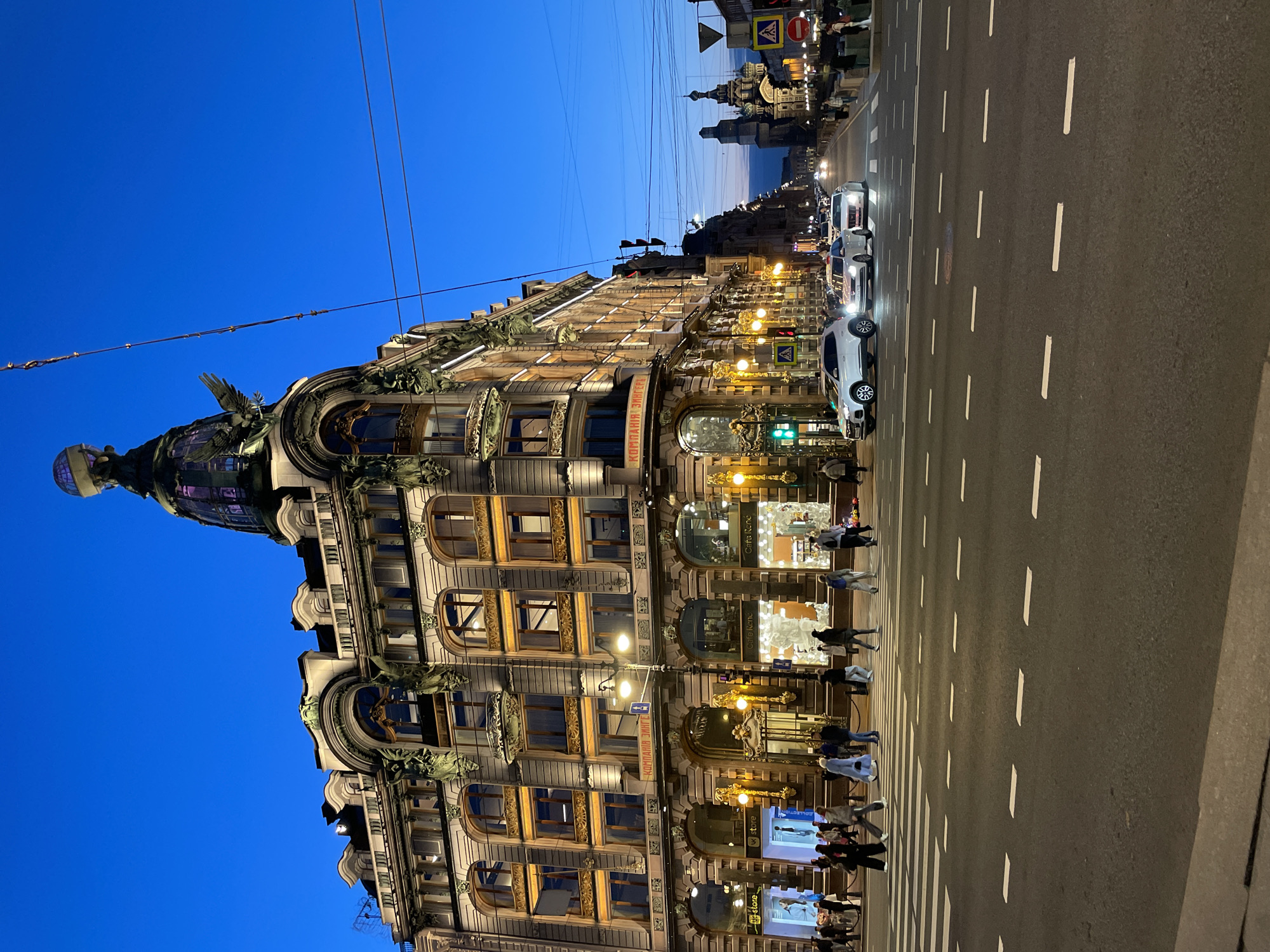 This evening image captures a vibrant street scene in Saint Petersburg, Russia, centered on the grand Zinger House (Dom Knigi) on the left under a deep blue twilight sky. The multi-story building, an iconic example of Art Nouveau architecture, is brightly illuminated by warm yellow lights, highlighting its ornate details, arched windows, and decorative sculptures, including a large bird, possibly an eagle, near the distinctive corner dome which is topped with a globe.
A wide asphalt street occupies the right side of the frame, featuring white dashed lane markings and a prominent crosswalk where pedestrians are seen. Several cars, mostly white, with illuminated headlights, are present on the road, suggesting ongoing traffic. Numerous people are visible walking on the sidewalk in front of the illuminated building and crossing the street.
In the distance, further down the street, other illuminated buildings and structures can be observed, including the distinctive domes of a church, likely the Church of the Savior on Spilled Blood. Overhead power lines traverse the sky. Visible text on the Zinger House includes "КОМПАНИЯ ЗИНГЕРЪ" (Singer Company) on a sign, and storefronts display "Cafe Rene", "W Store", and "COLLECTION". The photograph conveys a bustling urban atmosphere during the blue hour, characterized by historic architecture and commercial activity.