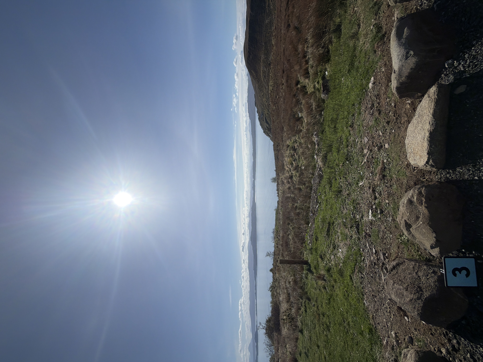 The image shows a landscape in Portree, United Kingdom, with a bright sun illuminating the sky on the left. A body of water, likely a loch or bay, stretches horizontally across the middle of the frame, separating the sky from a landmass. The landmass on the right side of the image consists of grassy terrain with brown, dry patches, suggesting late winter or early spring. Several large rocks are positioned near the bottom right corner. A small sign with the number "3" is visible near the rocks. The scene suggests a hiking trail overlooking the water.