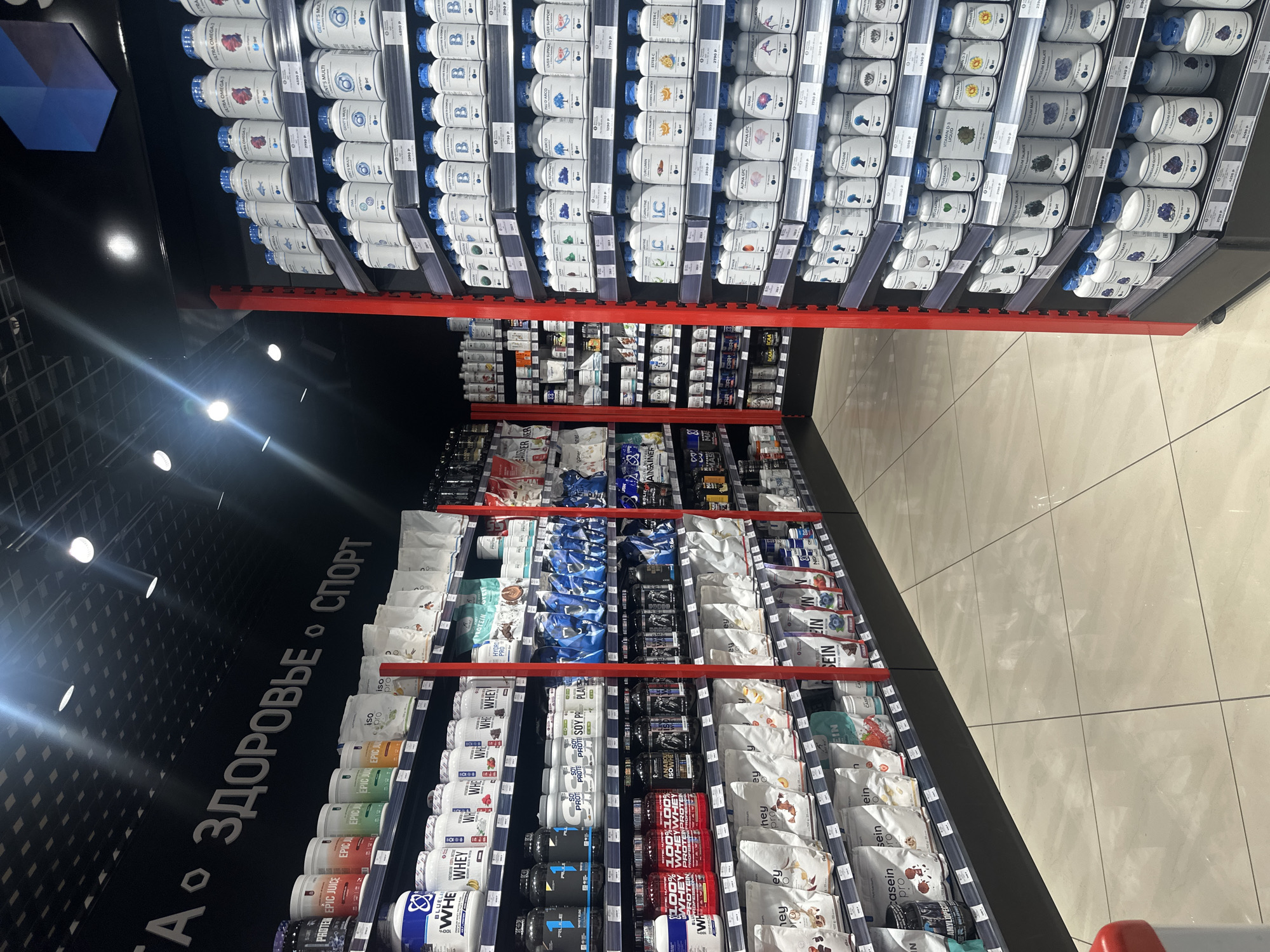 The image depicts the interior of a retail store specializing in sports nutrition and supplements, likely located in Petropavlovsk-Kamchatsky, Russia, as indicated by the signage and pricing conventions. Dominating the scene are shelves filled with numerous products in various containers: large tubs of protein powder, smaller bottles of pills and capsules, and foil-sealed bags. The products are organized by type and brand, with clear labeling in Russian. The store appears well-lit, with overhead lighting casting reflections on the tiled floor and the glossy surfaces of some product containers. The overall impression is one of a clean, organized, and well-stocked establishment catering to fitness enthusiasts.