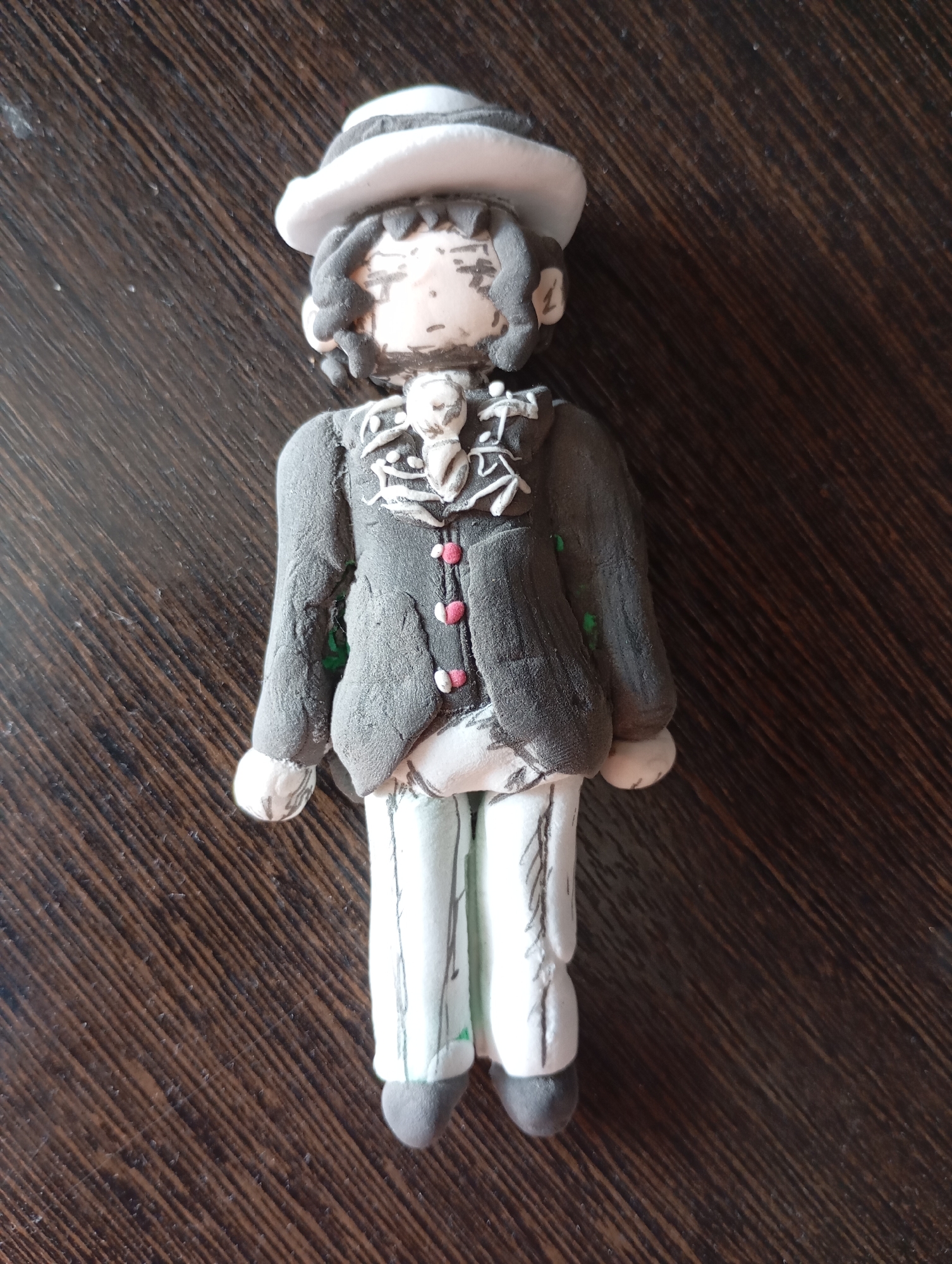 A handmade figurine of a person wearing a hat, jacket, and pants is laid out on a dark, textured wooden surface. The figurine has a pale complexion with black hair styled in curls. It wears a white and gray striped hat, a dark gray jacket with pink buttons, white pants, and dark gray shoes. The jacket has intricate white detailing around the collar and chest area. The background is a wooden table with diagonal grain patterns. The scene is lit from above, casting subtle shadows. The location in the city of Льниха, Russia, is not discernible from the image.