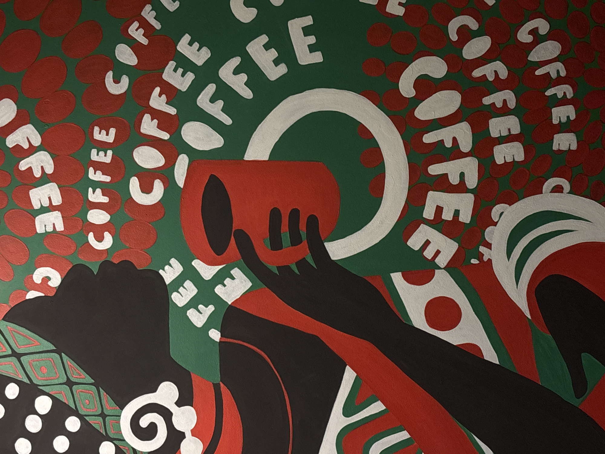 The image is an artwork depicting a person's profile in silhouette, holding a red coffee cup. The background is a deep green, with numerous red oval shapes representing coffee beans. The word "COFFEE" is written in white, stylized letters, repeated and arranged throughout the image in various orientations. The person's head is adorned with a patterned headdress in black, white, and green with red accents. They are also wearing a red garment with green and white accents. The artwork is likely a painting or a digitally created illustration. There is no information about the location of this artwork in the city of Perm, Russia.