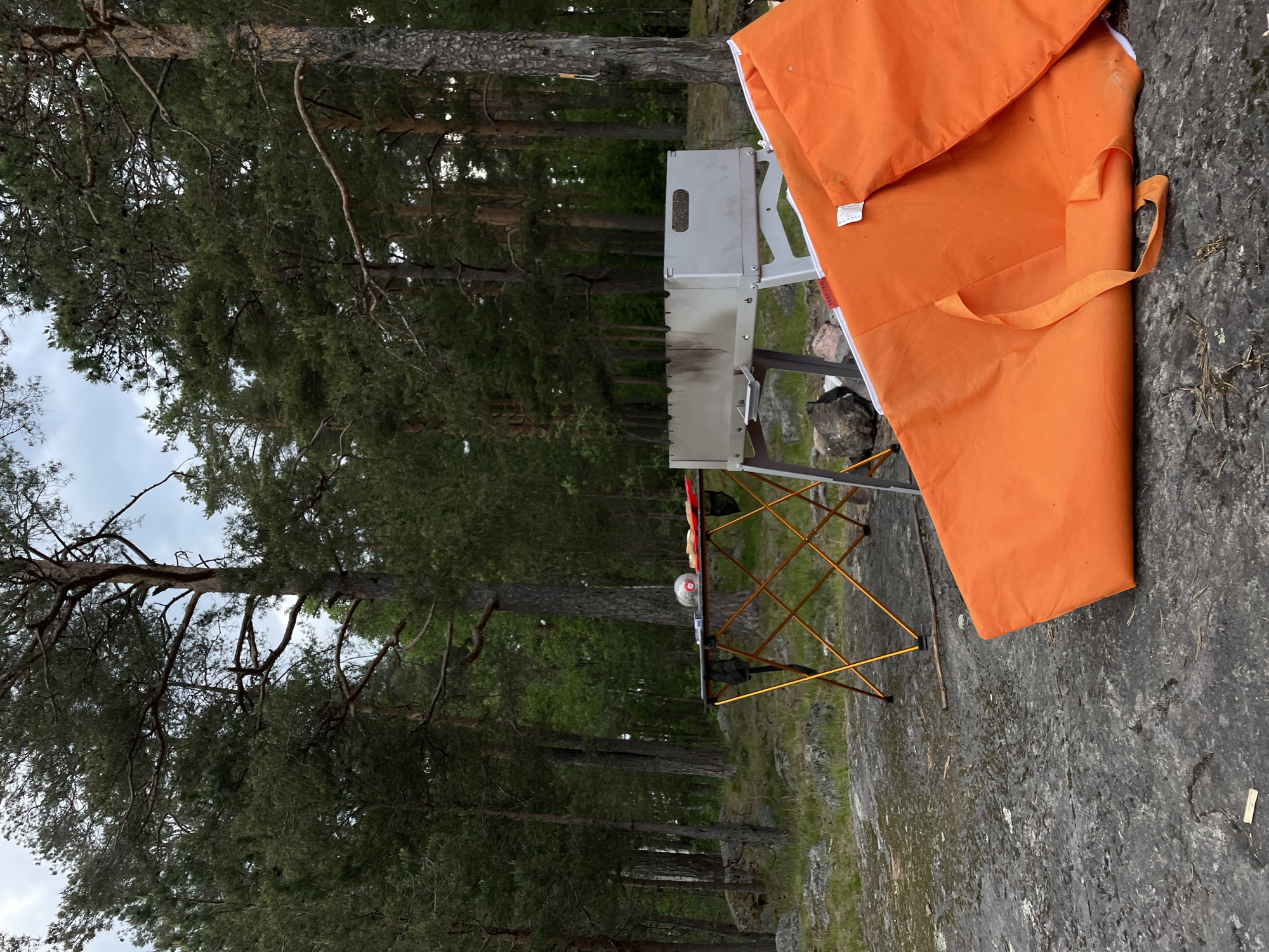 The image captures an outdoor recreational setting in Boitsovo, Russia, characterized by a forest environment and rocky ground under an overcast sky.
In the foreground, the ground consists of a large expanse of dark grey, rough-textured bedrock with some visible cracks and scattered small pieces of light-colored debris.
In the midground, an assemblage of portable camping equipment is present. To the left, a collapsible camping table with an orange-gold anodized aluminum X-frame supports a dark-colored tabletop. On the tabletop, a clear plastic bag containing a white spherical object (resembling a volleyball or similar ball) is positioned next to a red rectangular tray holding several small, light-colored, possibly bread-like, food items. A black mesh storage pouch hangs from the table's frame. Adjacent to the table on the right, a rectangular, metallic grey, portable barbecue grill (mangalnitsa) with side handles stands on its own folding support structure. The interior of the grill shows evidence of prior use, including charring and soot. In front of the grill, a large, vibrant orange fabric item, possibly a ground tarp or mat, is folded on the bedrock. It features a visible white border and an orange carrying strap, with a small white tag displaying partially obscured red text. A metal implement, possibly a skewer or barbecue fork, is seen hanging from the trunk of a pine tree directly behind the grill.
The background is composed of a dense coniferous forest, primarily populated by tall pine trees with dark green needles and rough, grey-brown bark. The forest floor recedes into the distance, showing an uneven topography of rocks, soil, and sparse undergrowth. The sky overhead is uniformly light grey, indicating overcast conditions. No individuals are visible within the scene, which appears to be a temporary outdoor cooking or picnic site.