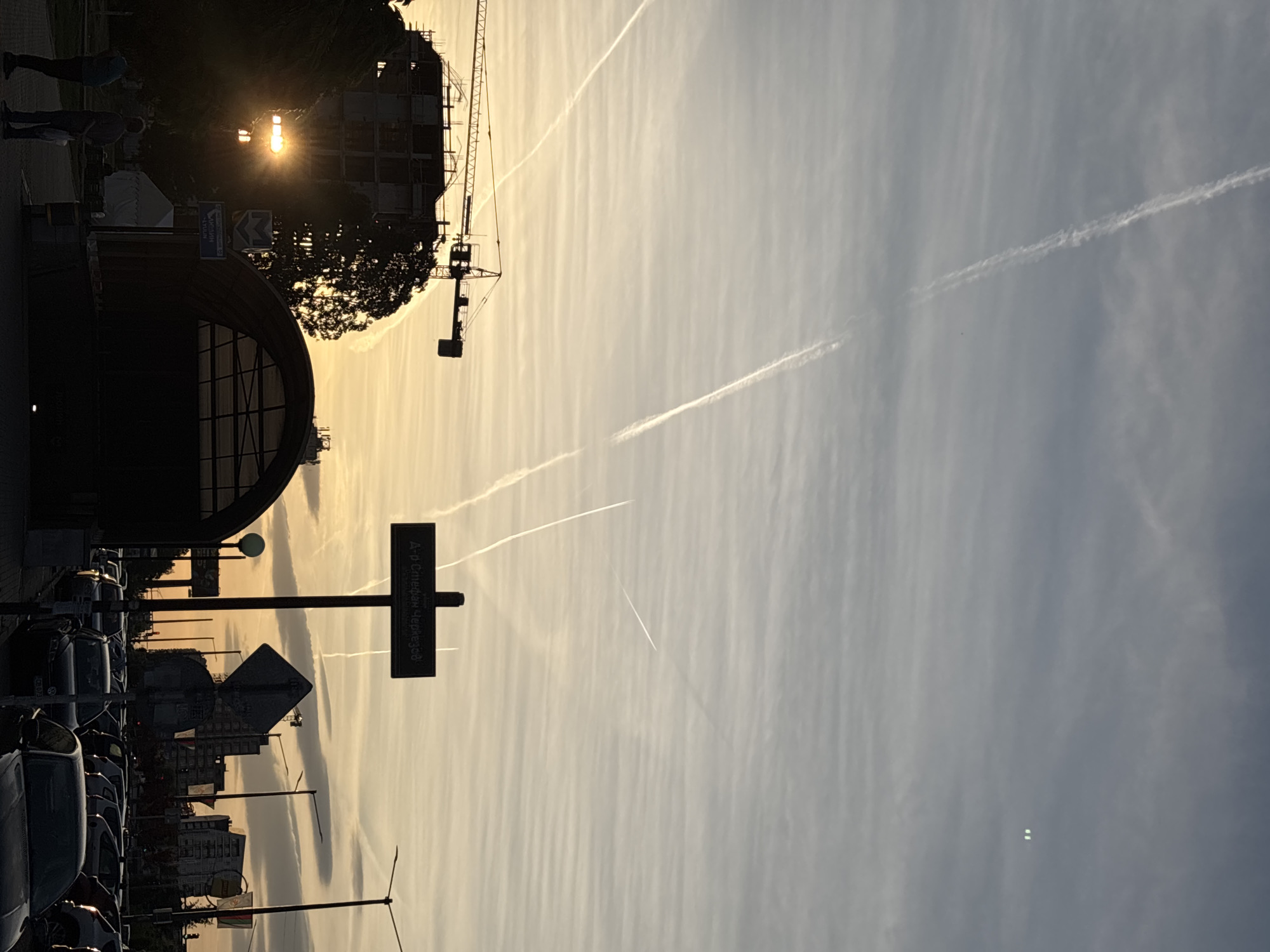The image shows an urban street scene in Sofia, Bulgaria, taken during sunset or sunrise. The sky is filled with linear clouds and contrails from airplanes.
A signpost indicates "А. Ст. Стефан Черкезов" (A. St. Stefan Cherkezov) as the street name. Several parked cars line the left side of the frame. A building with an arched entrance is visible on the left, and construction with a crane can be seen further in the background, silhouetted against the light. Some people are standing near the entrance of the building on the left. A street light stands near the signpost.