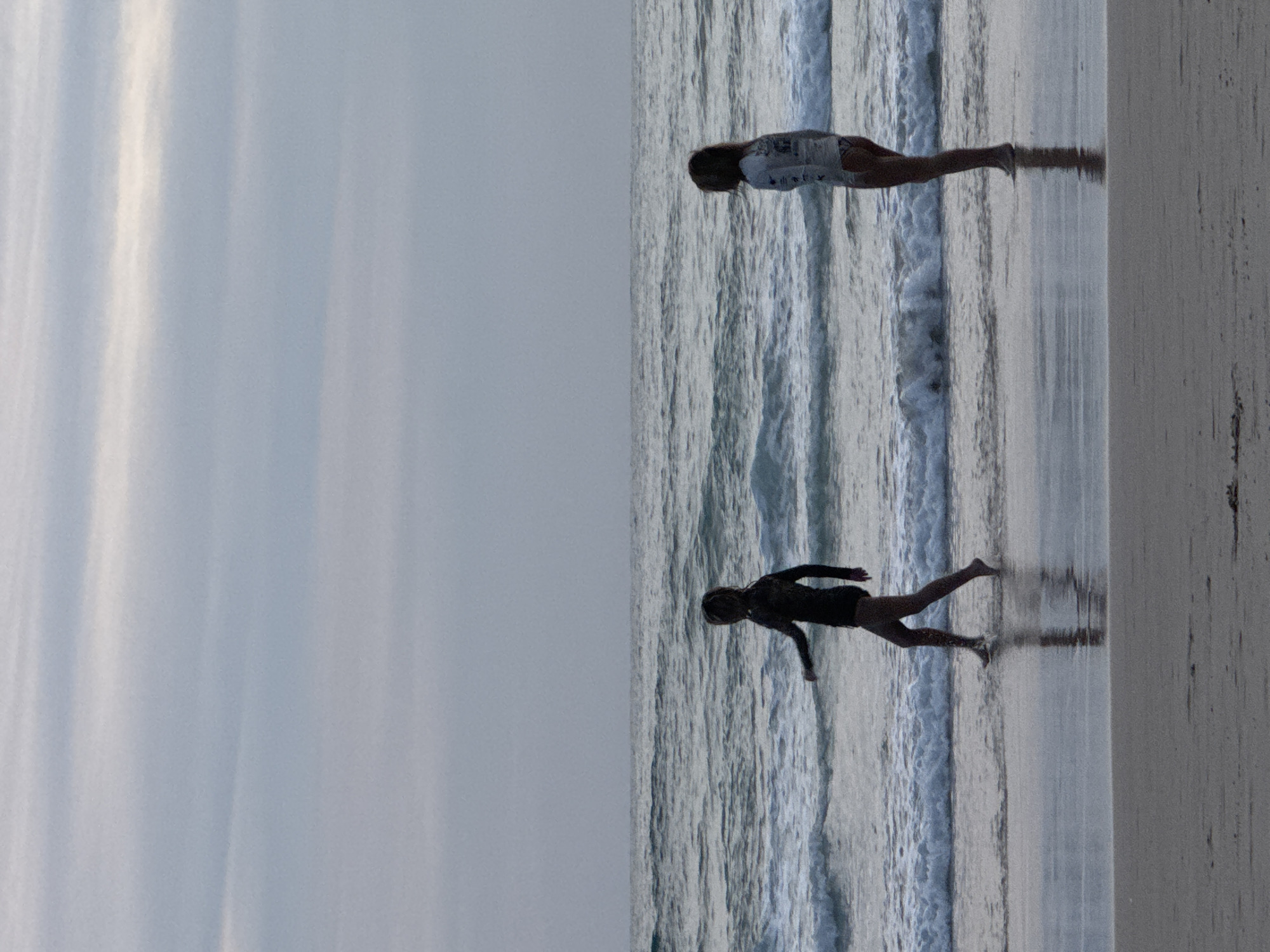 Two figures are walking on a wet sandy beach near the ocean, with waves gently lapping at the shore. The sky is overcast with streaks of light clouds, and the overall lighting suggests either early morning or late afternoon. The person on the left is wearing a dark, full-body wetsuit and is mid-stride, with their left arm extended slightly forward. The person on the right is wearing a lighter-colored long-sleeved rash guard and patterned bikini bottoms, and they are standing facing slightly away from the camera, looking out towards the ocean. The water is a deep blue-grey, and the wet sand reflects the light from the sky. The scene is tranquil, capturing a moment of quiet activity by the sea. Based on the prompt, this scene is set in Karon, Thailand.