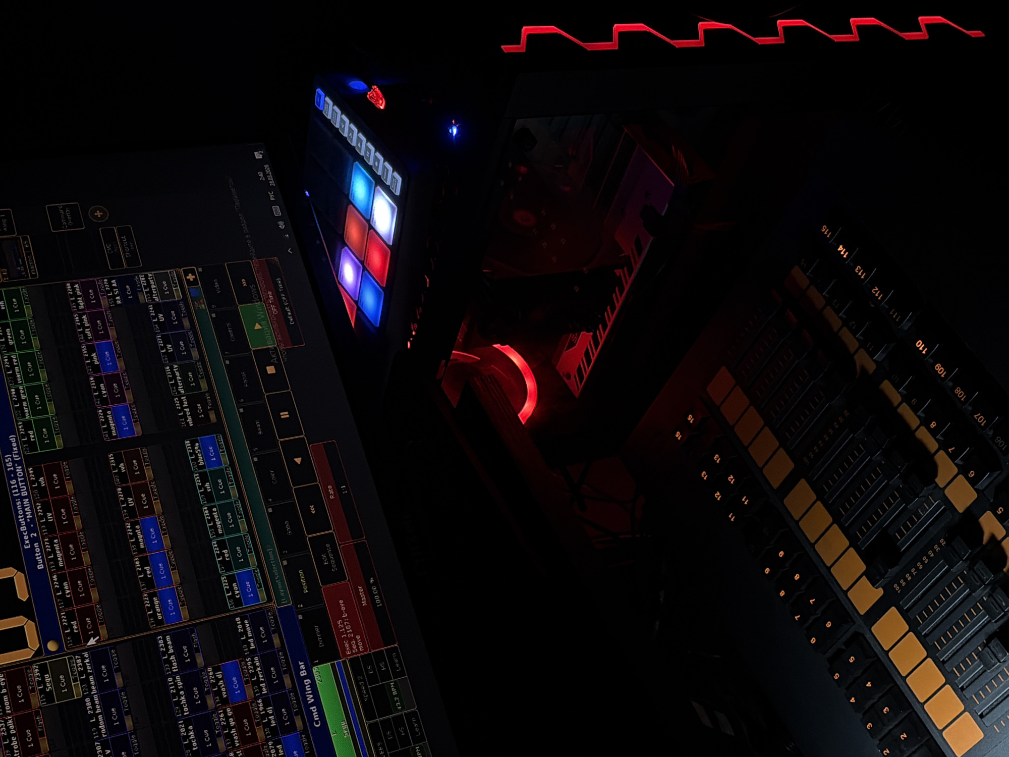 An interior control room within an entertainment venue or production facility in Moscow, Russia, is depicted in low light, illuminated by the operational glow of electronic equipment. A large monitor in the upper left displays professional lighting control software with a dark interface, showing multicolored text labels such as "red," "cyan," "magenta," "UV," and numerical sequences, alongside a Windows activation prompt and a system timestamp indicating "2:40 28.03.2020." Below the monitor, a compact digital control pad features six prominent square buttons illuminated in blue, red, and white, accompanied by smaller white-lit rectangular buttons and a small red emblem. Partially visible beneath this pad is a computer tower emitting a red internal glow from a fan and components, with a small blue indicator light on its top surface. In the foreground, a professional lighting or sound control console is visible, featuring numerous vertical faders with white markings and an array of orange-backlit square buttons and numerical indicators (e.g., 1-15, 105-115). A distinct vertical element with a continuous zigzagging red LED light strip frames the right side of the image. No individuals are present; the scene suggests an active, professional technical setup.