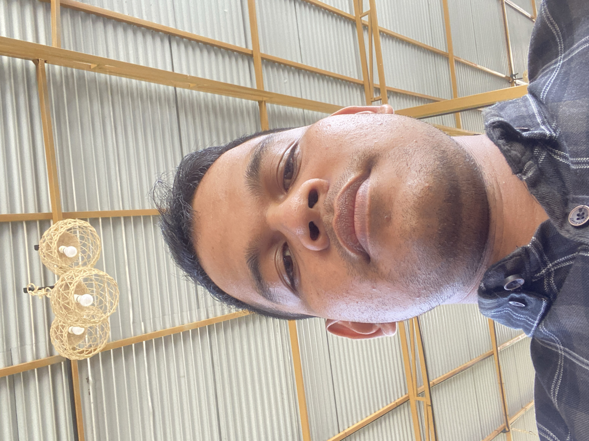 A low-angle, close-up selfie shows a person's face and upper torso. The person has dark hair, brown eyes, and a neutral expression. They are wearing a dark, plaid button-up shirt. The background consists of a corrugated metal ceiling with wooden beams and three woven pendant light fixtures. The lighting suggests an indoor or covered outdoor location. The provided information states this scene is located in Aluepiet, Indonesia.
