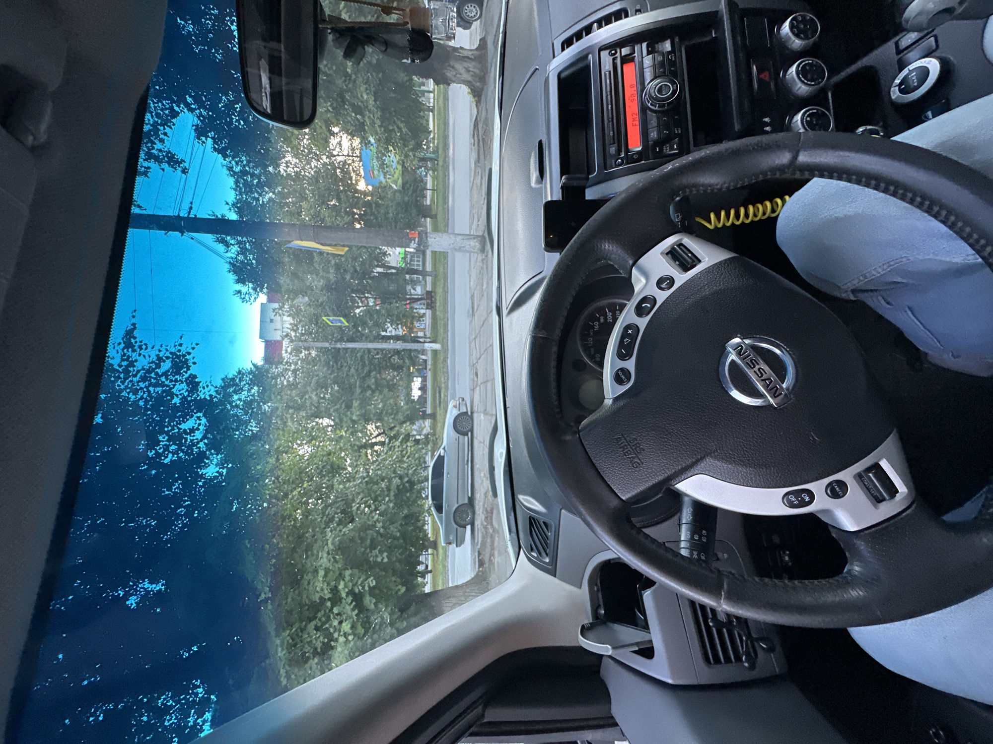 The image provides an interior view of a car, looking out through the windshield at a street scene. The perspective suggests the photo was taken by the driver. The car's dashboard is visible in the foreground, including the steering wheel with a Nissan logo, the instrument cluster, and the radio, which displays "FM2 94.8". A person's legs, clad in light blue jeans, are visible in the lower portion of the frame, resting on the driver's side.
Outside the car, a street is lined with trees and a sidewalk. A silver sedan is parked on the side of the road. A traffic light and a pedestrian crossing sign are visible in the distance, indicating a junction. A yellow and blue flag, likely the Ukrainian flag, is attached to a utility pole. Buildings can be seen beyond the trees. The sky is a bright blue with some dappled light filtering through the trees, suggesting it is daytime. The overall environment appears to be an urban or suburban setting. The presence of the Ukrainian flag strongly suggests the location is in Ukraine. Based on the information, it is highly probable that the city is Kamianets-Podilskyi, Ukraine.