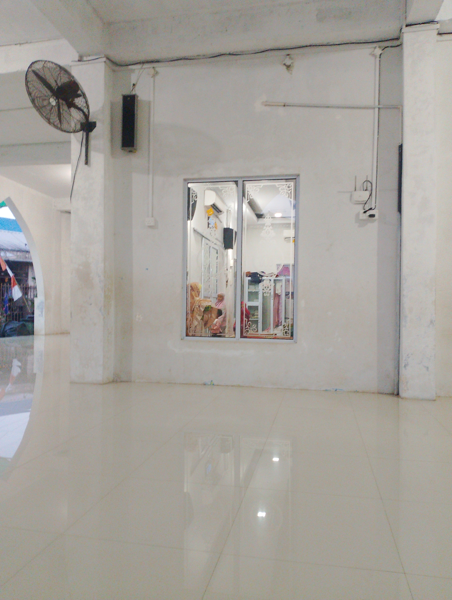 The image shows the interior of a building with white walls and a tiled floor. A large, black oscillating fan is mounted on the left wall, near the ceiling. A rectangular black speaker is mounted on the wall to the right of the fan. A double-paned window is centrally located in the wall. Through the window, two individuals are visible. One individual, wearing a pink garment, appears to be seated. The other individual is partially obscured and appears to be holding a light-colored object. Interior shelving units are visible behind them. Electrical conduits and a router are mounted on the wall to the right of the window. To the left of the window, an arched doorway leads to an exterior view, where a fence and flags are visible. The overall scene suggests an indoor community space or commercial area.