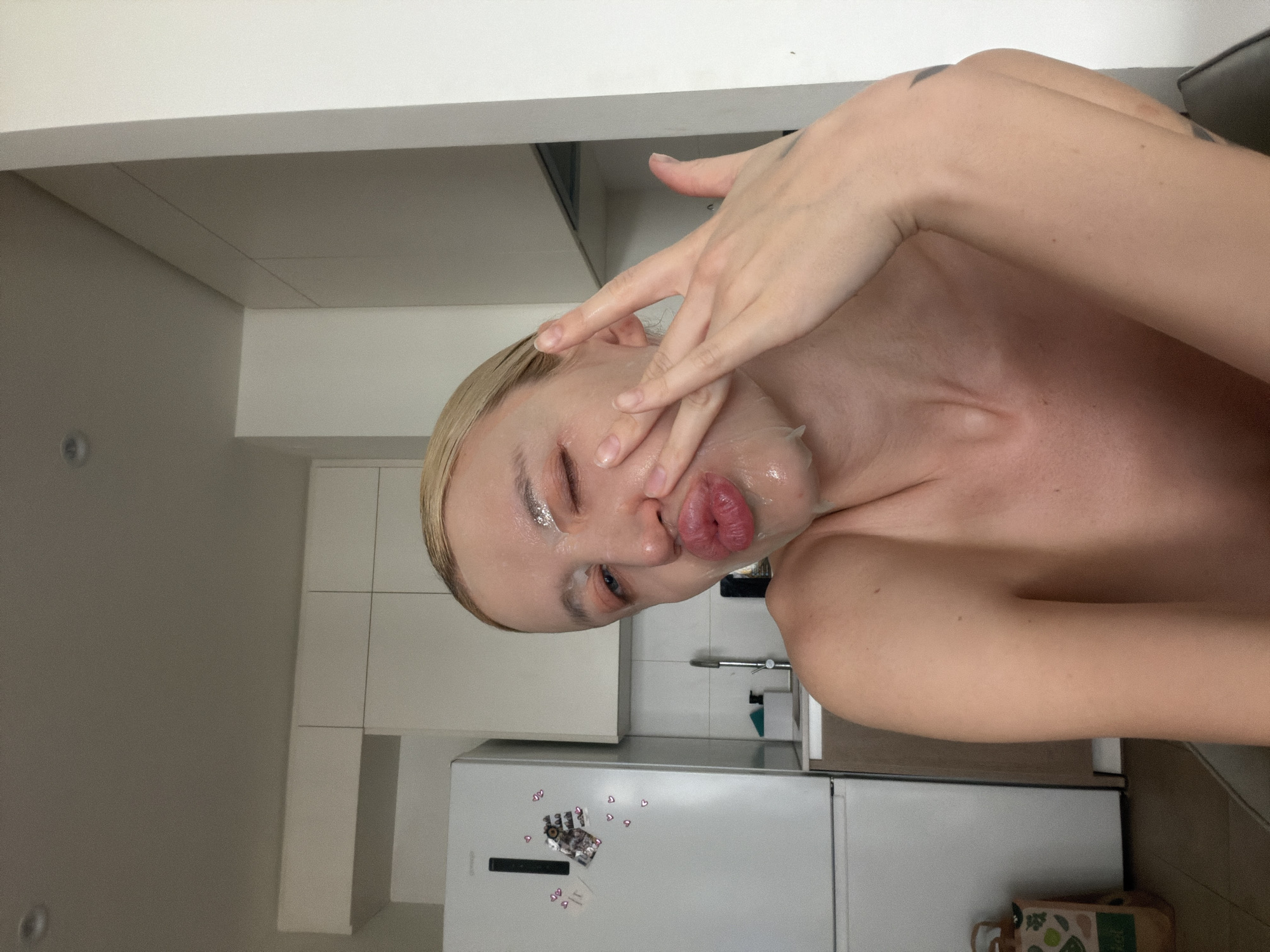 A shirtless person with short blonde hair and a translucent facial mask is posing for a photo in a kitchen. The person is winking with their left eye and puckering their lips while holding their hand up to their face. The kitchen has white cabinets and a refrigerator with magnets and a picture. A sink and countertop are visible in the background. The location is presumed to be an indoor residential space. The city of Belgrade, Serbia is not discernible from the visual data.