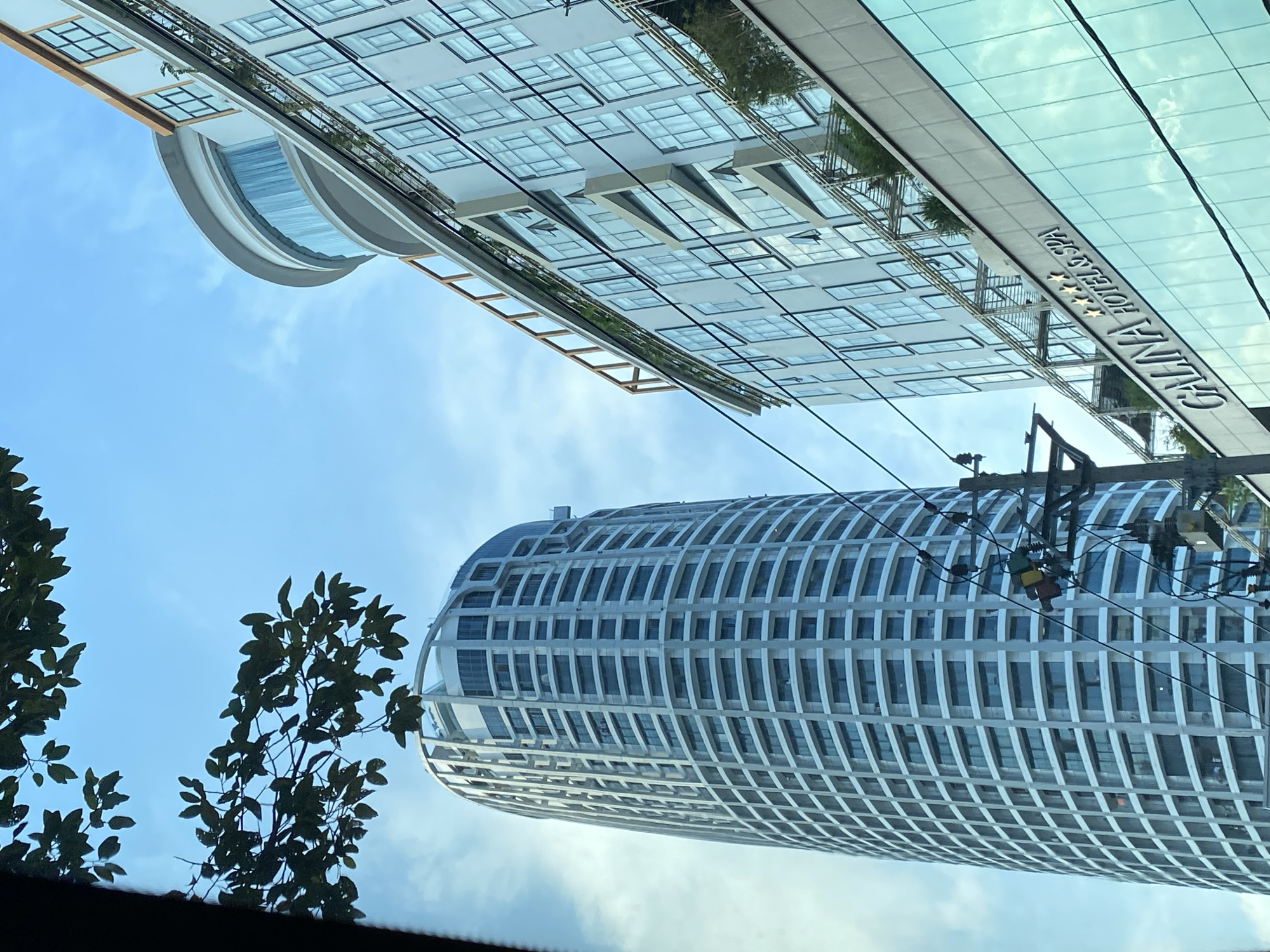The image depicts an exterior view of two modern buildings in Nha Trang Ward, Vietnam, under a bright sky with some clouds. On the left, a tall, cylindrical skyscraper with a white facade and numerous windows stands prominently. Its structure features vertical white beams that divide the facade into sections. On the right, a multi-story building with a glass facade, displaying a sign that reads "GALINA HOTEL & SPA," is angled towards the viewer. This building has a rounded section with large windows at its upper part, and several levels feature balconies with greenery. Overhead, dark green leaves from a tree partially obscure the sky in the upper left corner. Electrical wires and a utility pole are visible in the foreground, positioned between the viewer and the buildings. The scene appears to be captured from a street-level perspective, possibly from a moving vehicle.