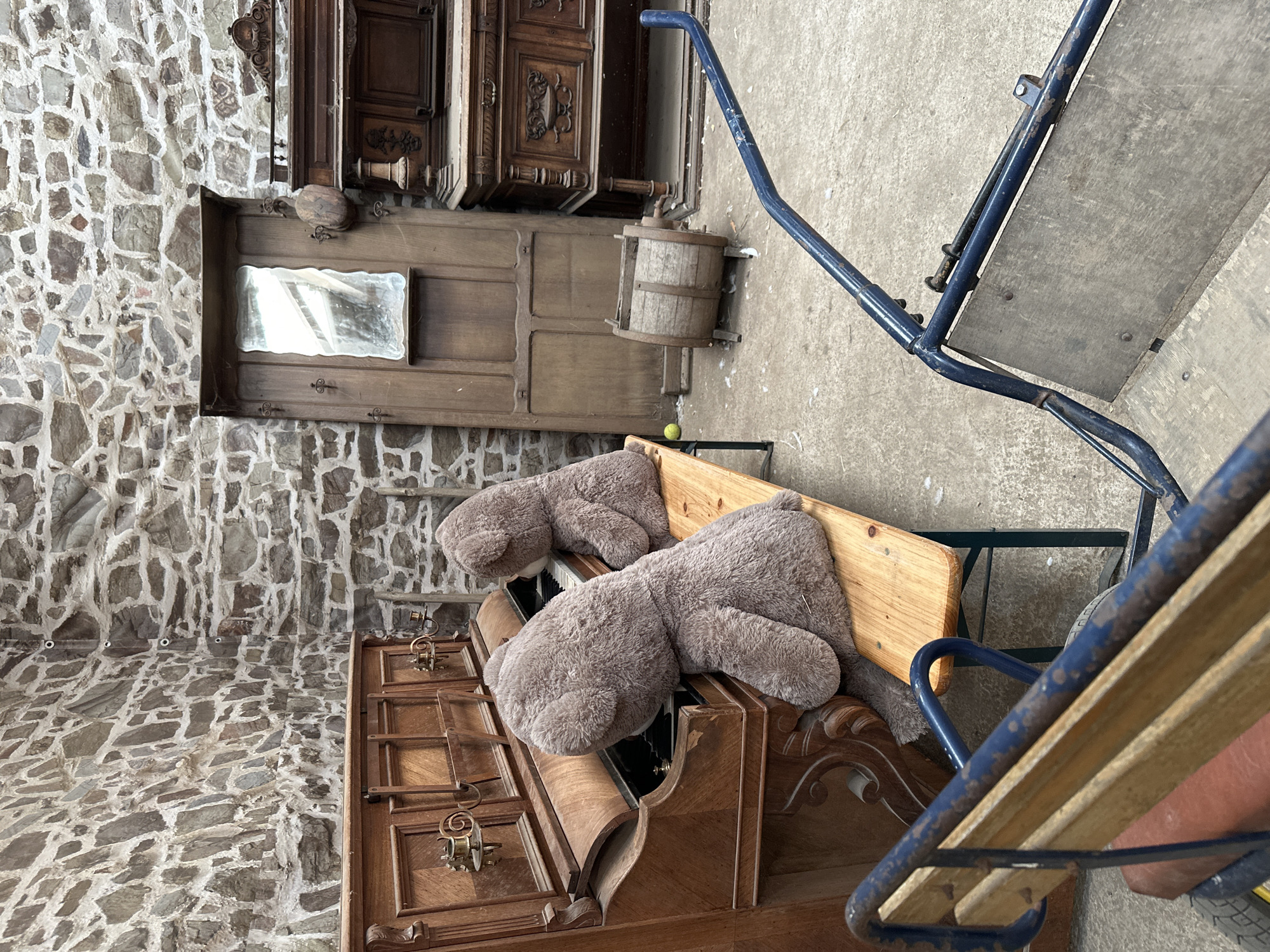The image depicts an indoor scene with a stone wall as the backdrop. In the foreground, a vintage wooden piano sits open, with two large, plush teddy bears seated on a wooden bench in front of it, appearing to play the instrument. Behind the piano, a tall wooden cabinet with a mirror and coat hooks stands against the wall. To the right of the cabinet are two ornate wooden furniture pieces, likely antique dressers or sideboards. In the middle ground, a small, wooden barrel-like object rests on the concrete floor. A blue metal cart with wooden components is partially visible in the lower right corner. The overall setting suggests a storage area or an antique shop. The image location is Gonneville-la-Mallet, France.