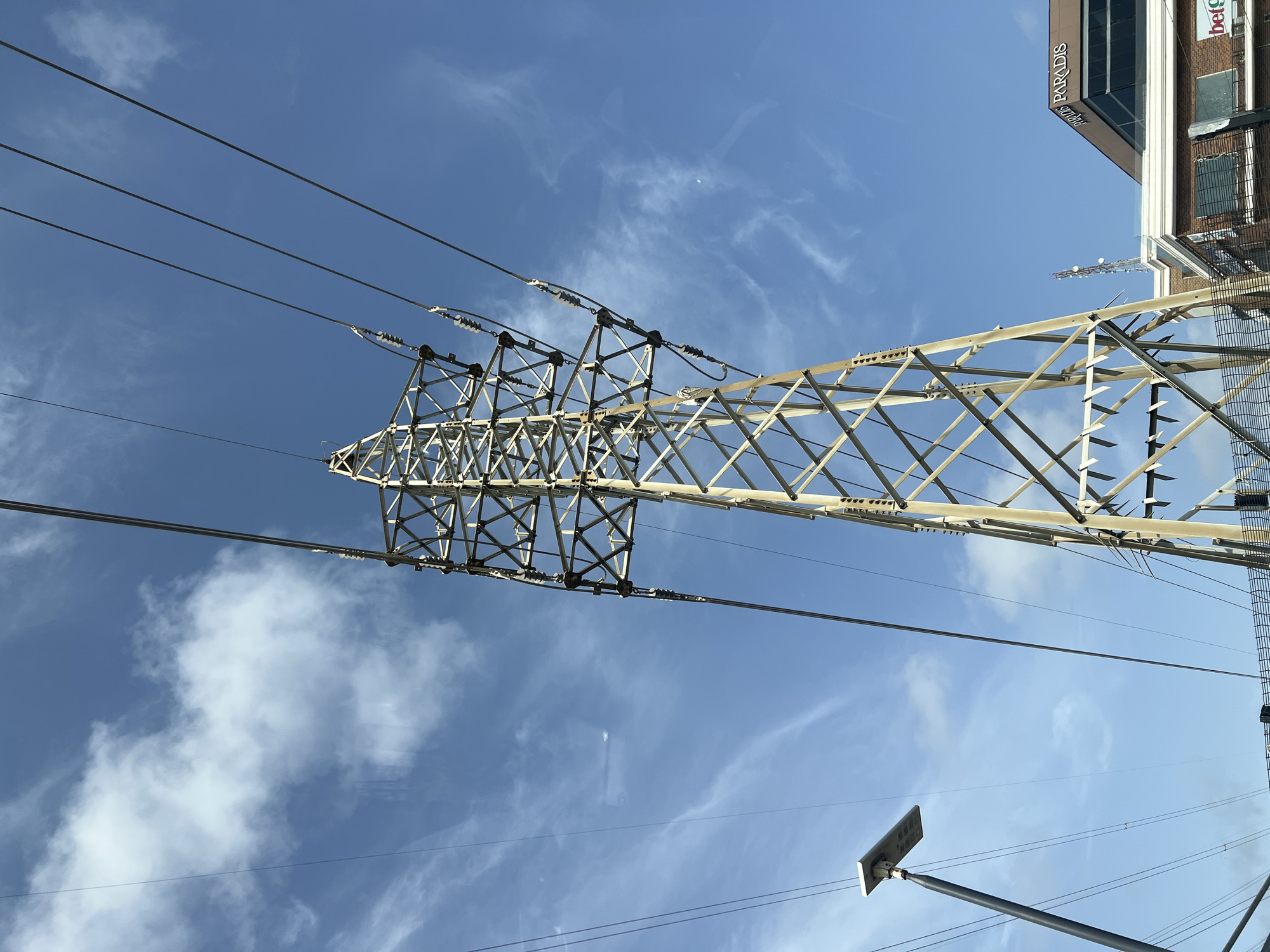 The image shows a low-angle view of an electrical transmission tower in Alaguntan, Nigeria. The tower is constructed of light-colored metal lattice. Multiple high-voltage power lines extend from the tower across a blue sky with scattered clouds. A building with signage visible, including "PARADIS" and "bet9ja" is partially visible in the top right corner. A street light is also present in the lower portion of the image, with wires running from it.
