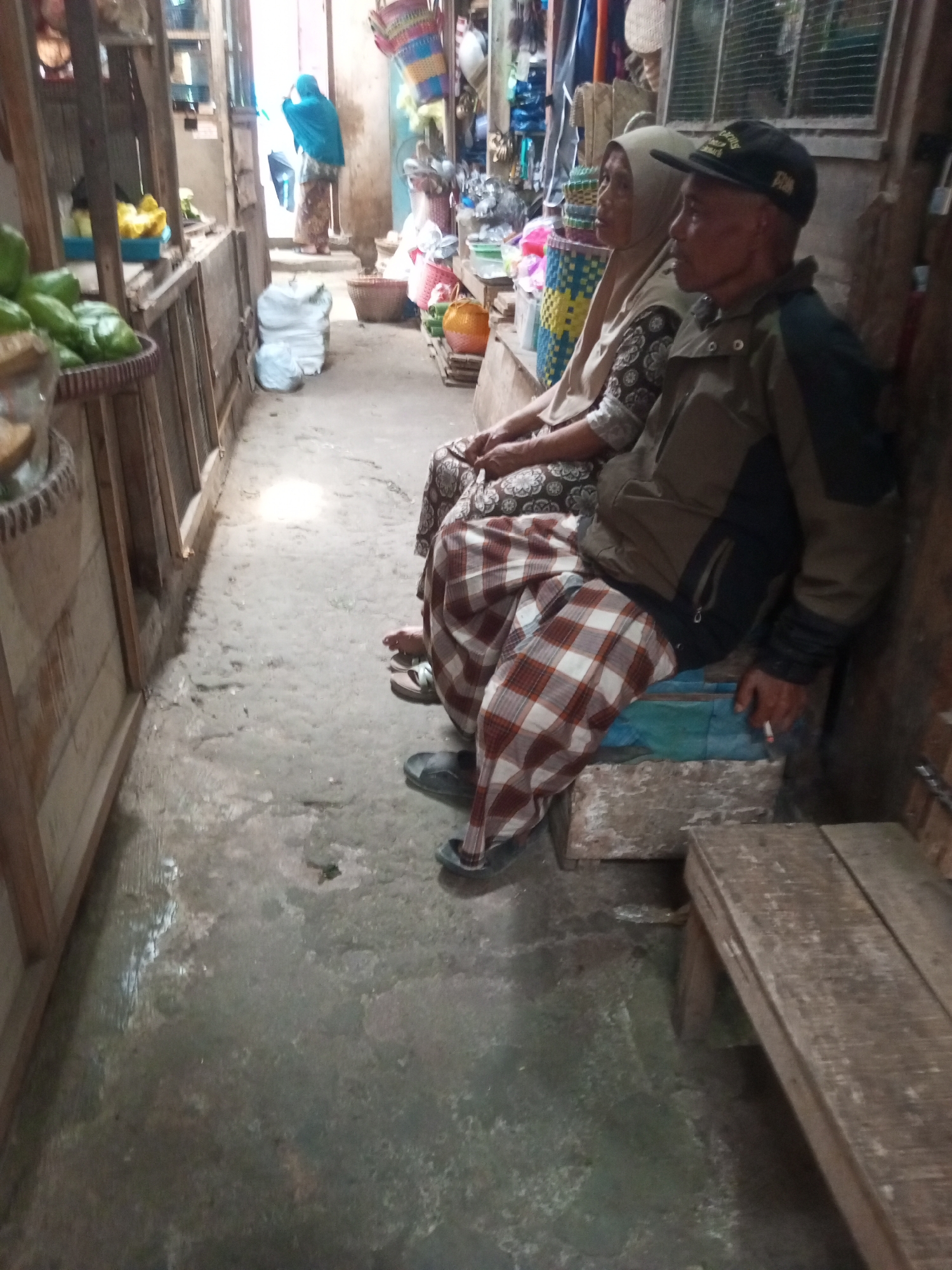 In Kalibeber, Indonesia, an elderly couple sits on a narrow, dirt-floored alleyway market. The man, wearing a dark baseball cap and a khaki jacket over a patterned shirt, smokes a cigarette. His lower half is clad in a red and white checkered sarong. Beside him, a woman in a light-colored hijab and a brown and white patterned dress sits with her hands in her lap. They are positioned on makeshift seats constructed from concrete blocks and a blue tarp. The background reveals market stalls laden with goods, including colorful woven baskets and sacks of produce, hinting at the commercial activity of the locale. The overall scene depicts a moment of rest amidst the bustling environment of a traditional Indonesian market.