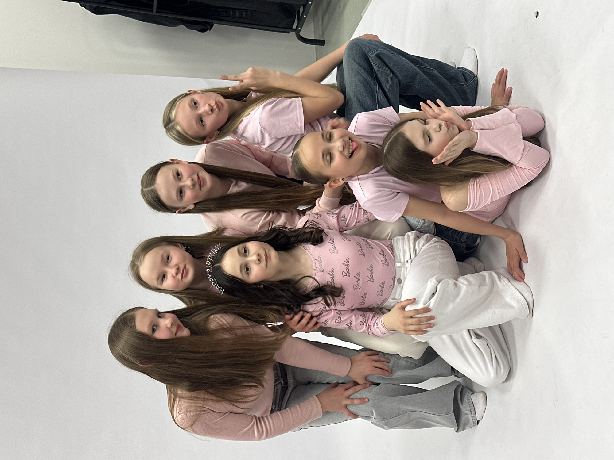 A group of seven young girls are posing for a photo in what appears to be a studio setting. They are arranged in a line, stacked on top of each other, wearing predominantly pink tops and various shades of jeans or white pants. The background is a plain white backdrop, suggesting a professional photo shoot environment.
The girls are making different poses and expressions, with some smiling, sticking their tongues out, making peace signs, or closing their eyes. One of the girls is wearing a "birthday" tiara.
The scene seems to be set indoors with controlled lighting. The black equipment visible in the top-left corner suggests the presence of studio lighting and possibly other photography gear.
There is no information to indicate that this photo was taken in Votkinsk, Russia.