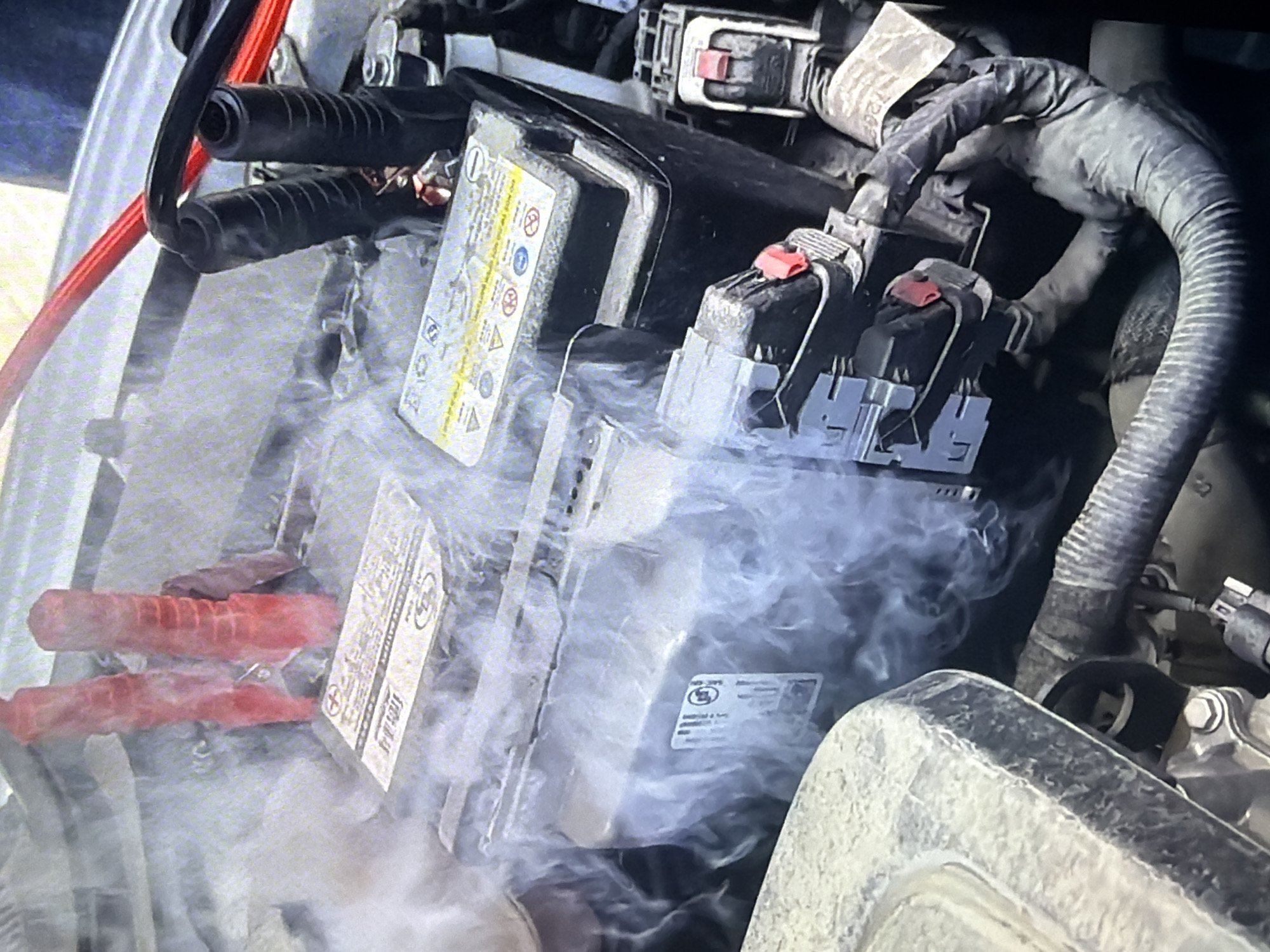 This image depicts a car battery in an engine bay, with visible smoke emanating from it. Red jumper cable clamps are attached to the battery terminals. The scene appears to be an outdoor setting, possibly a roadside in Bukuru, Nigeria, given the context of the request. The smoke suggests a potential malfunction or overheating of the battery. There are no people clearly visible in the image. The environment is somewhat dusty, with visible engine components and wiring.