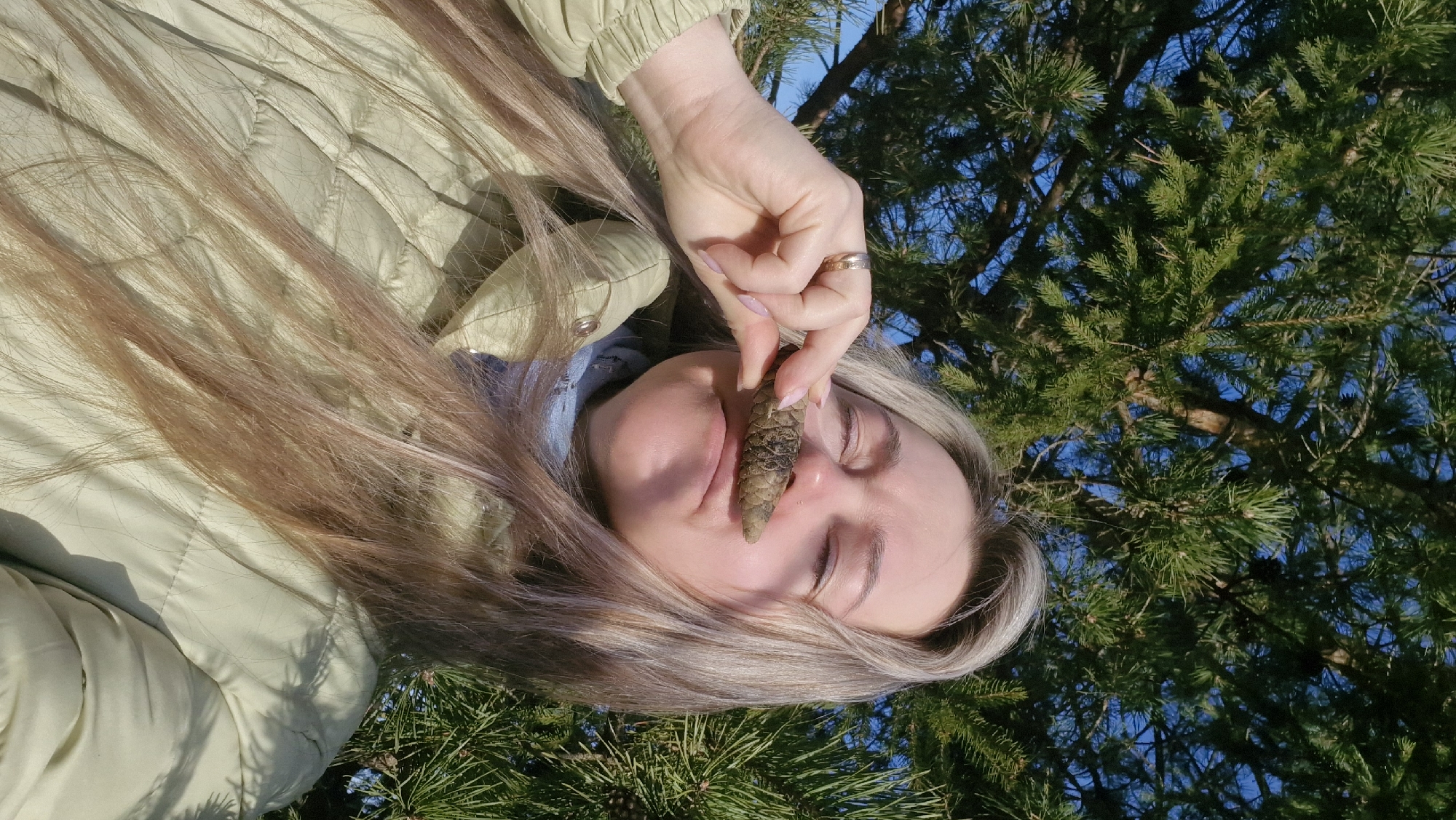 A woman with long, blonde hair is outdoors, likely in a wooded area or park. She is holding a pine cone between her fingers, positioning it near her nose. The background features pine trees and a blue sky. She is wearing a light-colored, quilted jacket. The picture seems to be taken at Otchor, Russia during a sunny day.