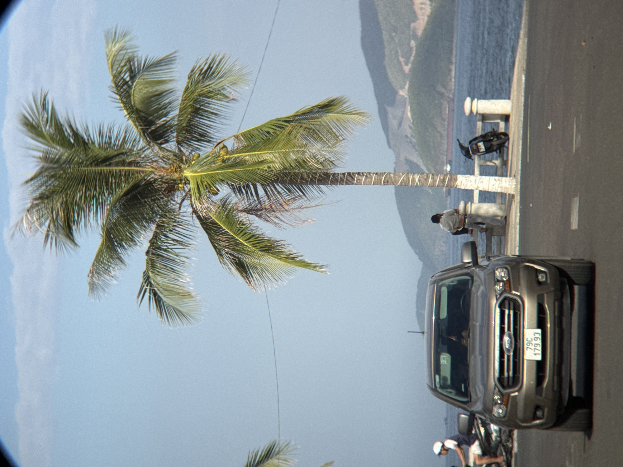 This image captures a street scene in Nha Trang, Vietnam, with a focus on a brown Ford Ranger pickup truck parked on a paved road. The truck's license plate reads "79C 179.93". To the left of the truck, a person wearing a white helmet and a blue vest rides a motorcycle. In the background, a tall palm tree dominates the frame, its fronds reaching towards a clear blue sky. Further in the distance, the blue ocean meets a lush green hillside, with a ship visible on the water. Two individuals are seated on a stone bench near a white stone railing, and a black motorcycle is parked behind them. The overall atmosphere suggests a coastal city environment with clear weather.