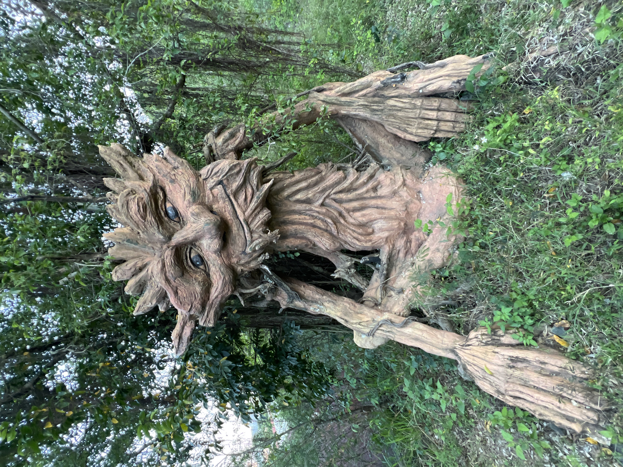An anthropomorphic sculpture, resembling a seated tree-like entity or "Ent," is positioned in a verdant outdoor setting within Nha Trang, Vietnam. The sculpture, crafted from material mimicking gnarled wood and bark, features a distinct face with deeply set eyes containing dark blue pupils. A prominent, textured nose and a beard-like formation of vertical bark-like strands are visible. Irregular, spiky protrusions extend from the top of the head. The body and robust limbs are contoured to simulate rough tree bark, extending to form large, root-like hands resting on the ground. The sculpture's coloration is a blend of earthen browns and reddish-browns, showing variations in texture and tone. It is surrounded by a dense layer of green grass and assorted small plants in the immediate foreground. The background is filled with abundant green trees, leafy foliage, and numerous hanging vines, creating a natural and lush environment. Diffused natural light illuminates the scene, casting soft shadows.