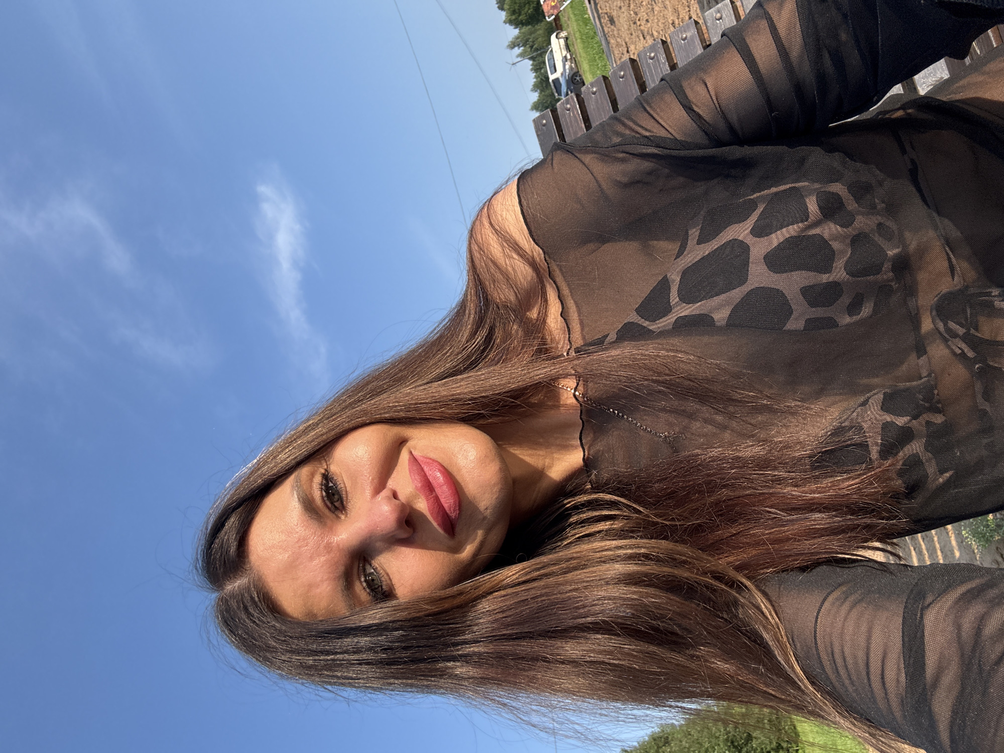 A woman with long, dark brown hair and makeup smiles at the camera outdoors. She is wearing a sheer black top with a leopard print bra underneath. The background shows a clear blue sky with a few wisps of clouds, trees, a wooden bench, and a parked car. The scene appears to be a park or recreational area, possibly in Prikamskii, Russia, given the surrounding vegetation and rustic wooden bench.