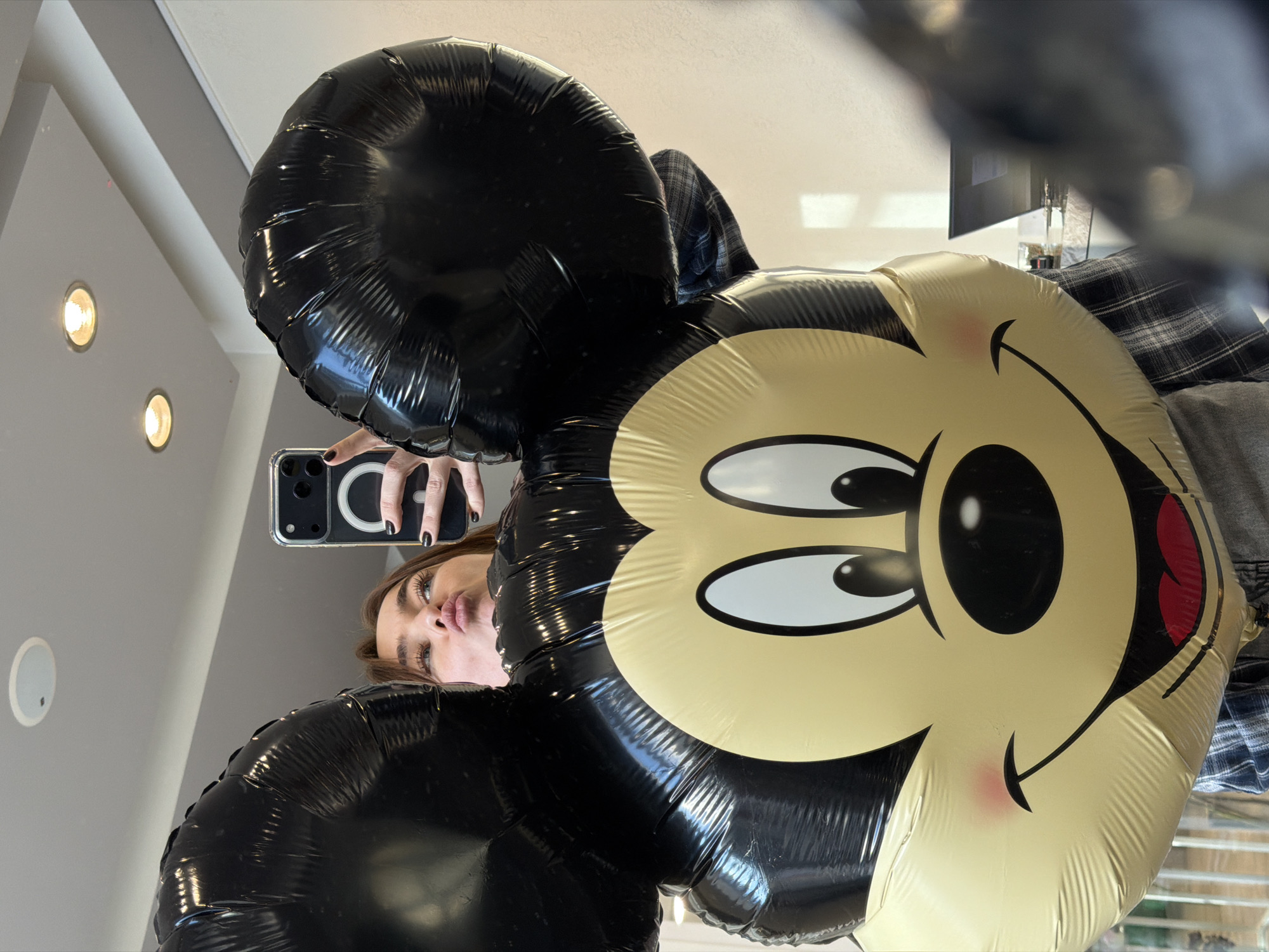 A woman with dark fingernails is holding a smartphone in her left hand, taking a selfie in front of a large Mickey Mouse balloon. The balloon is black and gold. The location appears to be indoors, with ceiling lights visible in the background. The city of Ust-Ilimsk, Russia cannot be determined from the visual information available.