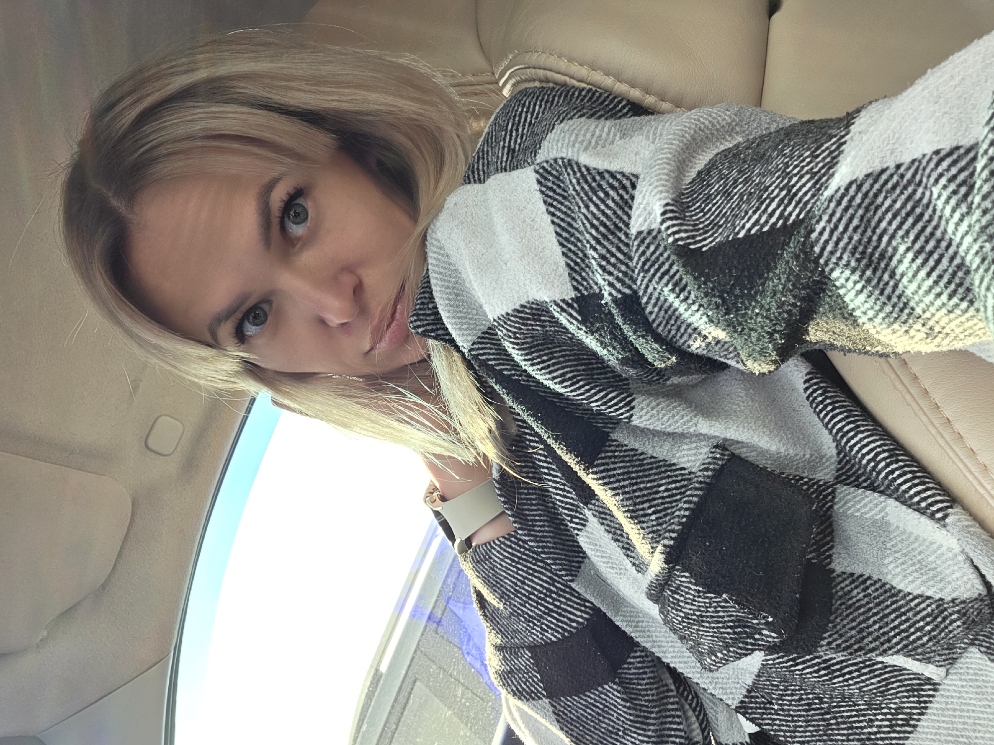 A blonde woman is sitting in the passenger seat of a car. She is wearing a black, white, and gray plaid shirt. The interior of the car is visible, with a beige leather seat and part of the headliner. Outside the car, the sky is bright and appears to be daytime. The image is a selfie, taken by the woman. No specific location in Chaikovsky, Russia, can be identified from the image.