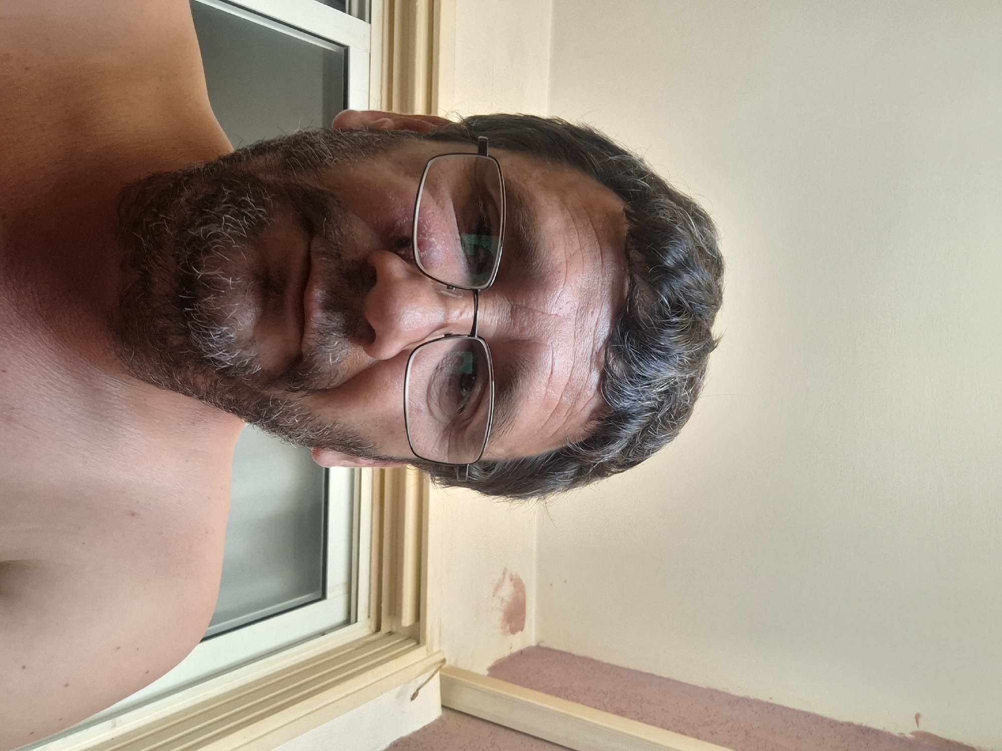 A close-up, low-angle selfie of a man wearing glasses, with a beard and short, dark, wavy hair. He has light-colored skin and is shirtless. The man is looking directly at the camera. Behind him, there is a window with a white frame and blinds. The ceiling is visible above, with some light-colored paint and a pinkish stain. The background wall on the right appears to be under construction or renovation. The image is undated and unlocated, but based on the context of other images provided, it is understood to be in Yerocham, Israel.
