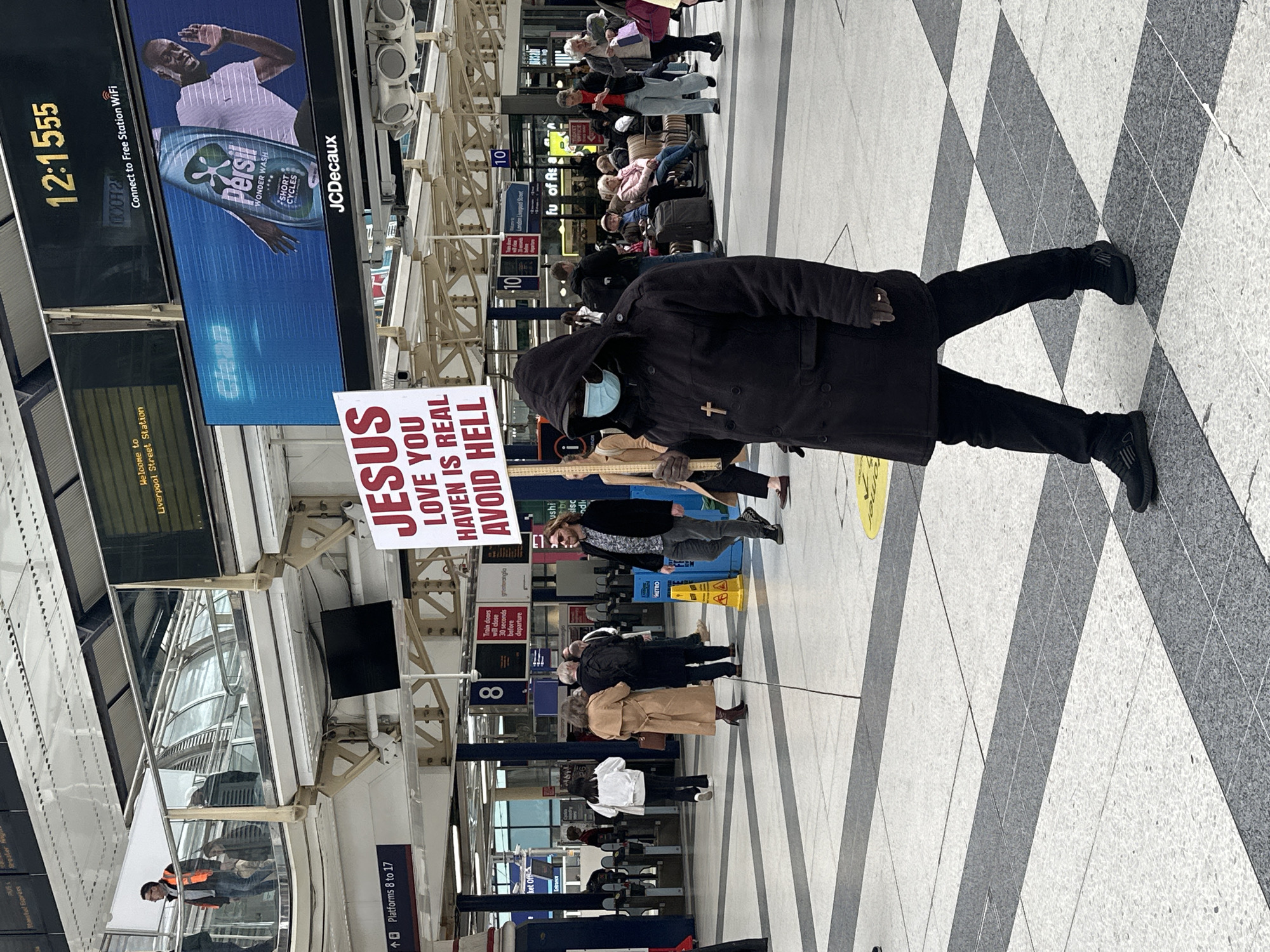 The image depicts a man holding a sign with the words "JESUS LOVE YOU, HAVEN IS REAL, AVOID HELL" in what appears to be Liverpool Street Station in London, United Kingdom. The man is wearing a dark coat with a cross on it, a hood, a blue mask and black shoes. The scene shows other people in the background, including passengers waiting for trains. Above the sign holder, there are digital advertising screens displaying time as well as commercial advertisements. The floor has a black and white patterned tile design. There are signs indicating platform numbers and station facilities.