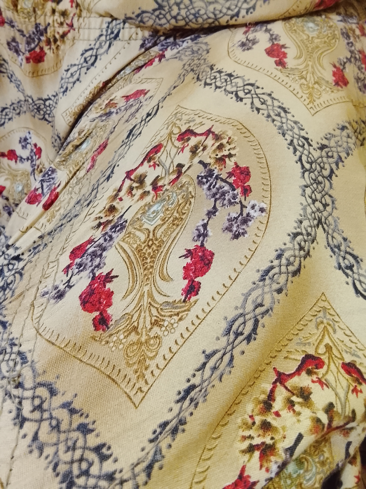 The image shows a close-up of patterned fabric. The fabric has a light beige background with repeating designs featuring floral motifs in red, purple, and brown. These motifs are contained within stylized, arched shapes outlined in a darker shade of beige. Separating these arched motifs are geometric patterns resembling intertwined ropes or chains in a bluish-gray color. The fabric is creased and folded, indicating it is not flat. No people or a specific location are discernible.