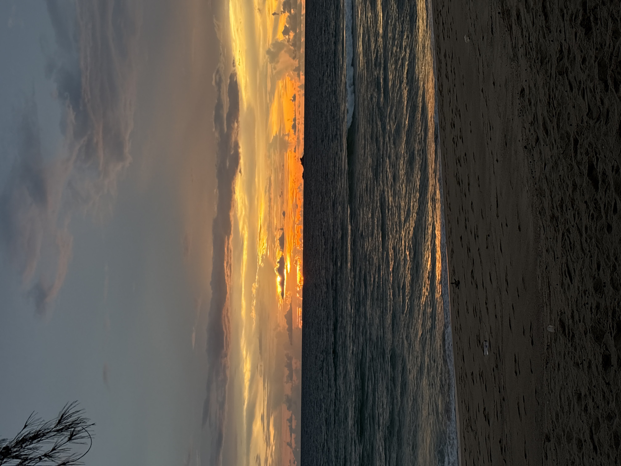 The image depicts a serene sunset over the Andaman Sea, likely near Choeng Thale, Thailand. The sky is a canvas of warm oranges, yellows, and muted grays as the sun dips below the horizon. Clouds are scattered across the sky, some illuminated by the sun's rays, creating a dramatic effect. The sea is a deep, calm blue with gentle waves lapping at the shore. A faint reflection of the sunset can be seen on the water's surface. In the distance, a small, dark silhouette of an island or landmass is visible on the horizon. The foreground is a sandy beach, sparsely dotted with footprints and a few pieces of debris, suggesting human presence. A branch of a dark-leaved tree hangs in the upper left corner of the frame, adding a touch of natural framing to the scene. The overall atmosphere is peaceful and tranquil, characteristic of a beach sunset.