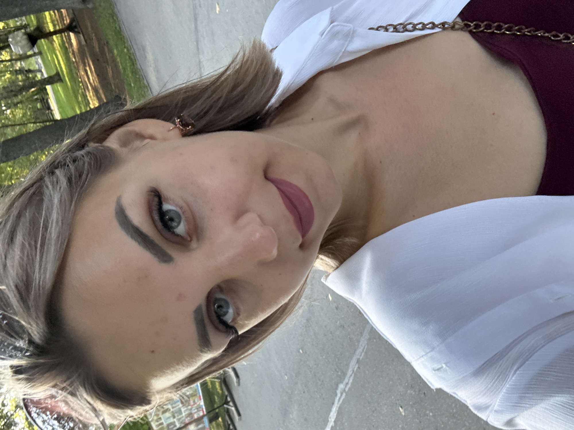 A young woman with blonde hair, blue eyes, and full lips is taking a selfie outdoors. She is wearing a white shirt over a burgundy top and a gold earring in her left ear. Her makeup includes dark eyeliner and mascara, and her lips are painted a matte pinkish-red color. The background shows a park with trees and a paved pathway. The woman's face is angled towards the camera, and she appears to be smiling slightly. The image was captured in Yuzhno-Sakhalinsk, Russia.