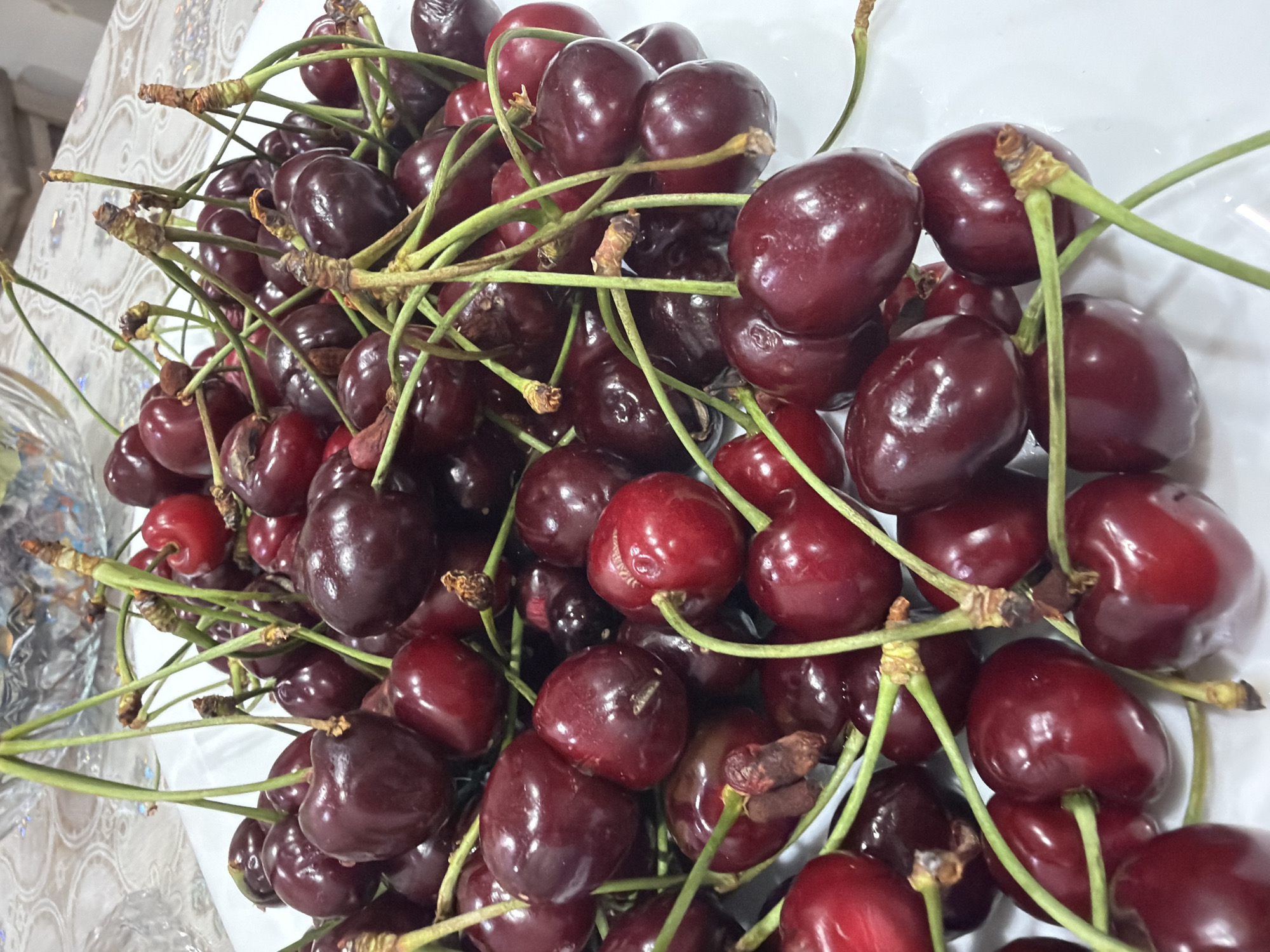 A close-up shot shows a pile of dark red cherries with green stems on a white plate. The cherries are glossy and appear to be fresh. The stems are thin and delicate, with some of them bearing small brown buds. In the background, a blurred patterned tablecloth and a partial view of a glass bowl are visible, suggesting an indoor setting. The lighting is soft and diffused, highlighting the texture and color of the cherries. The image does not provide any information about the city of Yerevan, Armenia, or any specific details about people, actions, or interactions.