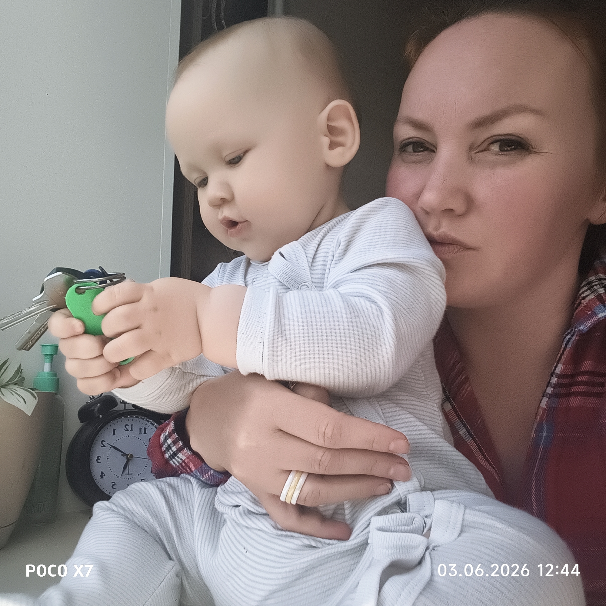 An indoor full-shot shows a woman holding a baby. The woman, with reddish-brown hair, is visible from the chest up, wearing a red and blue plaid shirt. Her expression is neutral, and she looks towards the viewer with one eye slightly squinted. She wears three rings on her left hand's ring finger: two white and one gold.
The baby, with fair skin and light hair, is held in the woman's arms, facing away from the viewer but looking down at an object in their hands. The baby wears a light-colored, striped long-sleeved outfit. In the baby's hands are a set of keys, including two silver keys and a bright green plastic keyfob.
The background behind the subjects is a light grey wall. To the left, an alarm clock with a black casing and a grey face with Roman numerals is partially visible, showing hands around 11:30 or 5:50. Next to the clock, a green-topped pump bottle and a small plant in a pot are also visible.
The image has a digital overlay in the bottom left corner that reads "POCO X7", indicating the device used. In the bottom right corner, the date and time "03.06.2026 12:44" are displayed. The scene captures a tender moment between a parent and child, with the baby engrossed in examining the keys.
