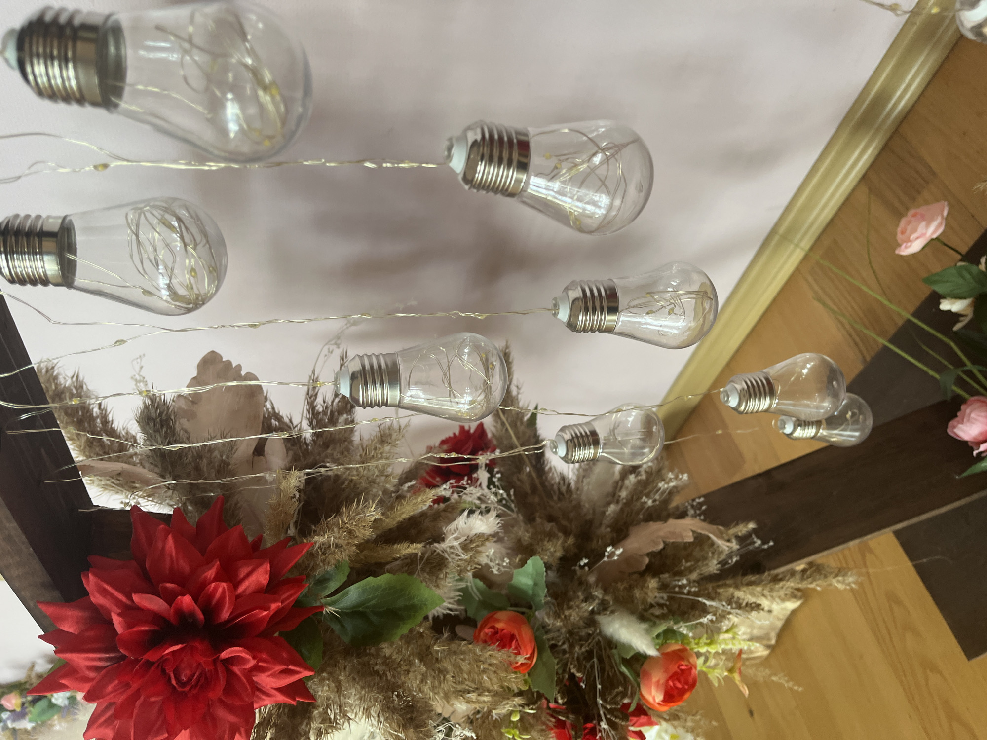 The image shows a decorative arrangement of artificial flowers and grasses with a string of decorative light bulbs. The light bulbs are clear glass in a vintage shape, each containing a small string of fairy lights. The flowers include a large red dahlia, several smaller red and orange roses, and white accents. The grasses are primarily brown and tan. The arrangement is set against a cream-colored ceiling and a wooden structure with varying shades of brown. The location is not identifiable as Saint Petersburg, Russia, based on the visual information provided.