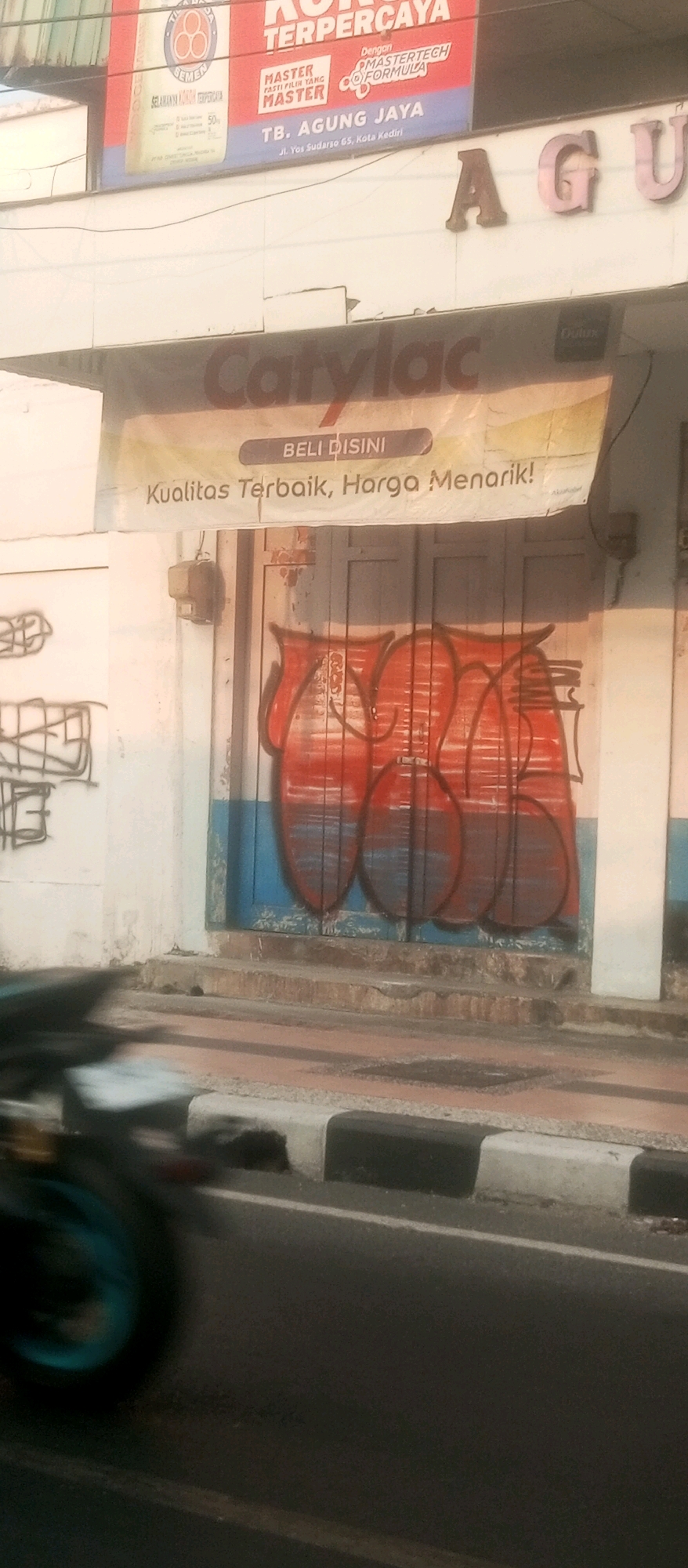 The image depicts a storefront in the city of Kediri, Indonesia. A building facade features a large sign with the text "AGU" visible, suggesting it may be part of a larger brand name or establishment. Below this, a banner reads "Carylac" and "BELI DISINI" (Buy Here) with the tagline "Kualitas Terbaik, Harga Menarik!" (Best Quality, Attractive Price!). The entrance to the storefront is a double door, painted blue and white, with red graffiti spray-painted across it in a bold, abstract style. To the left of the entrance, there is a white wall with black graffiti tags. A utility box is mounted on the wall to the right of the door. The storefront is situated on a street with a curb and sidewalk. In the foreground, a blurred motorcycle wheel is visible, indicating motion. The overall scene suggests a commercial area with signs of urban wear and tear, including peeling paint and graffiti.