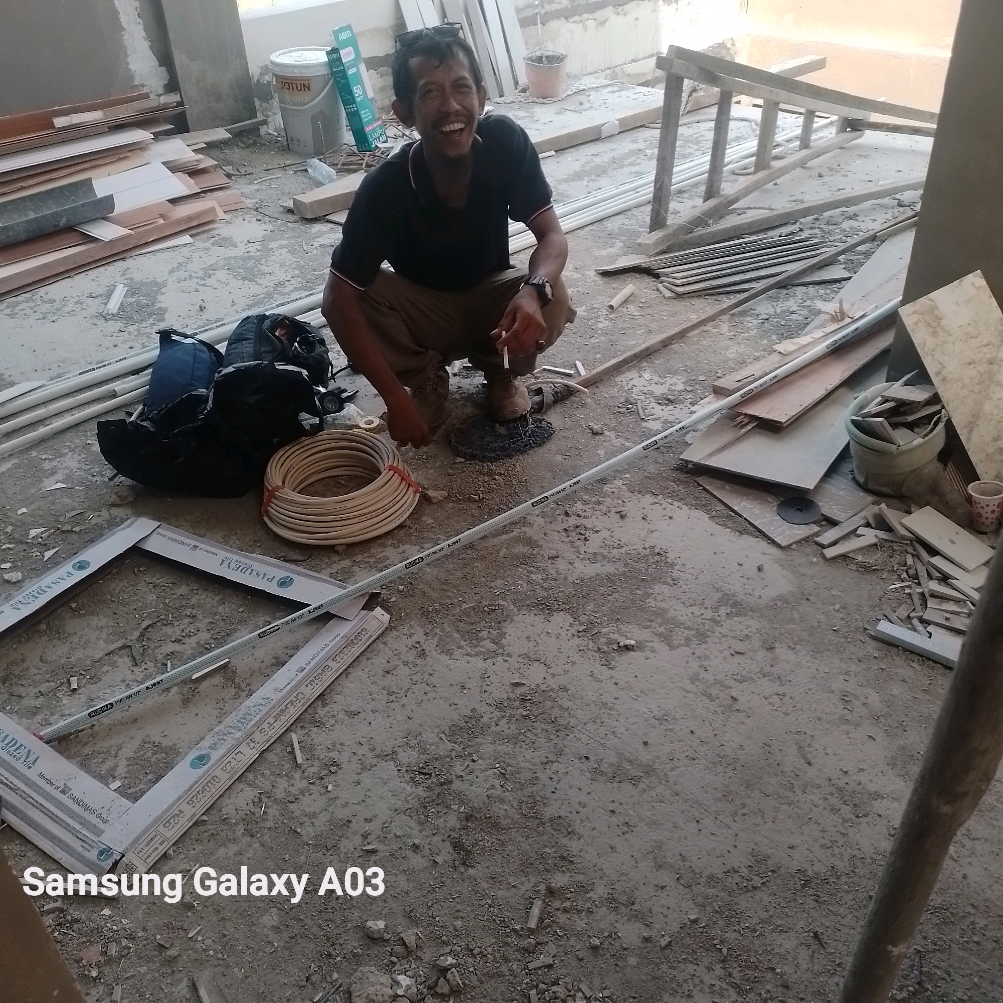 A man is crouching on a construction site in Kenanga, Indonesia, laughing. He is wearing a black t-shirt and khaki pants, and he holds a cigarette in his right hand. In front of him are several construction materials, including pipes, lumber, and bags. The ground is a mixture of dirt and debris. The scene appears to be an outdoor construction area, possibly a new building or renovation project. The image is captured with a Samsung Galaxy A03 phone.