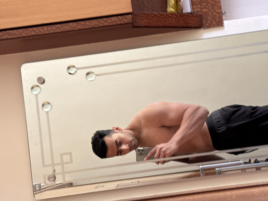 A person is visible in a mirror taking a photo of themself. The person is shirtless, wearing dark shorts. The mirror is attached to a surface or furniture, which can be seen at the top and bottom edges. The mirror's design includes rounded decorative elements. The photo captures the reflection of the person's head, torso, and part of their legs, along with the smartphone they are using to take the photo.